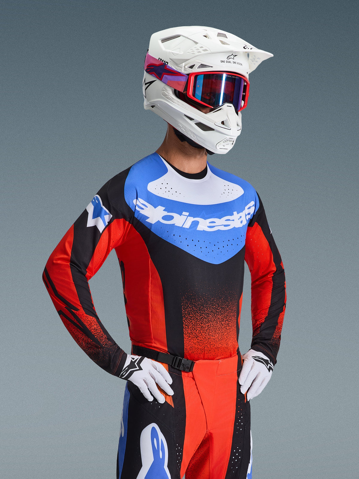 Pilota di motocross con completo Alpinestars Techstar Knif 2026, maglia blu con marchio bianco e maniche rosse, pantaloni neri e rossi coordinati con accenti blu, casco bianco con occhiali rossi, guanti bianchi e neri, abbigliamento professionale per motocross
