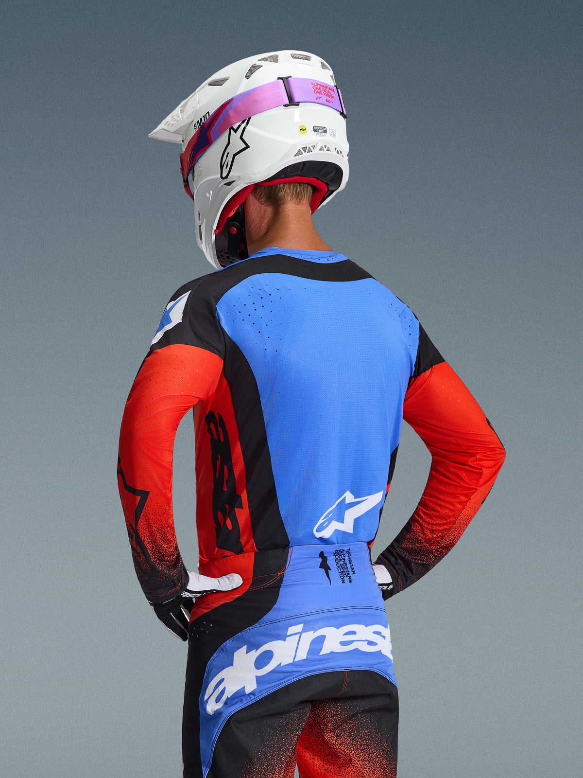 Pilota di motocross visto da dietro che indossa il completo Techstar Knif 2026, maglia blu con maniche rosse e accenti neri, pantaloni neri e blu con marchio Alpinestars bianco, casco bianco con elastico degli occhiali rosa, con traforature al laser per la ventilazione