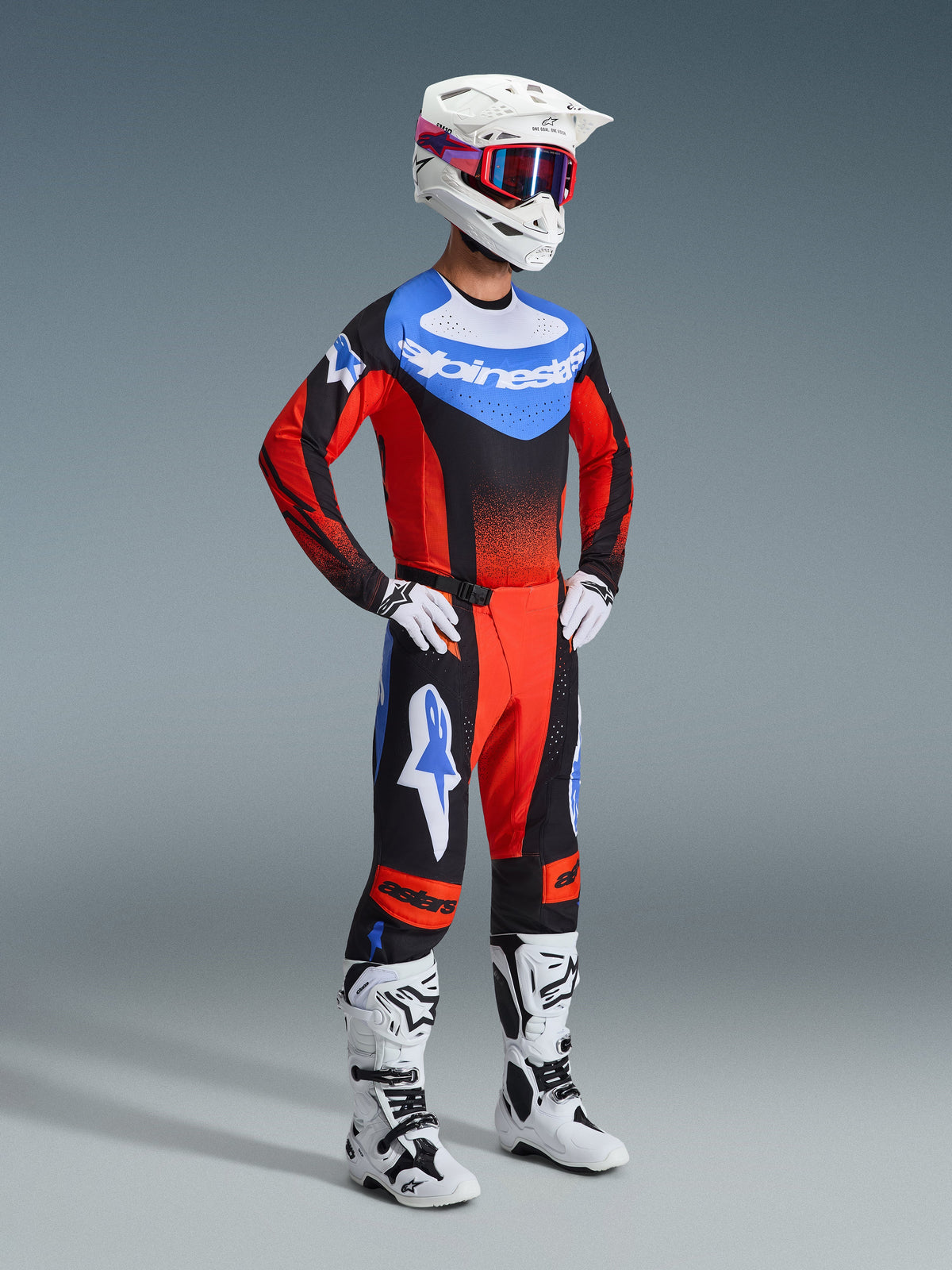Pilota di motocross con completo Alpinestars 2026 Techstar Knif, maglia con petto blu, logo bianco e maniche rosse sfumate di nero, pantaloni coordinati neri e rossi con dettagli a stella blu, casco bianco con occhiali rossi e blu, stivali bianchi e neri e guanti bianchi.