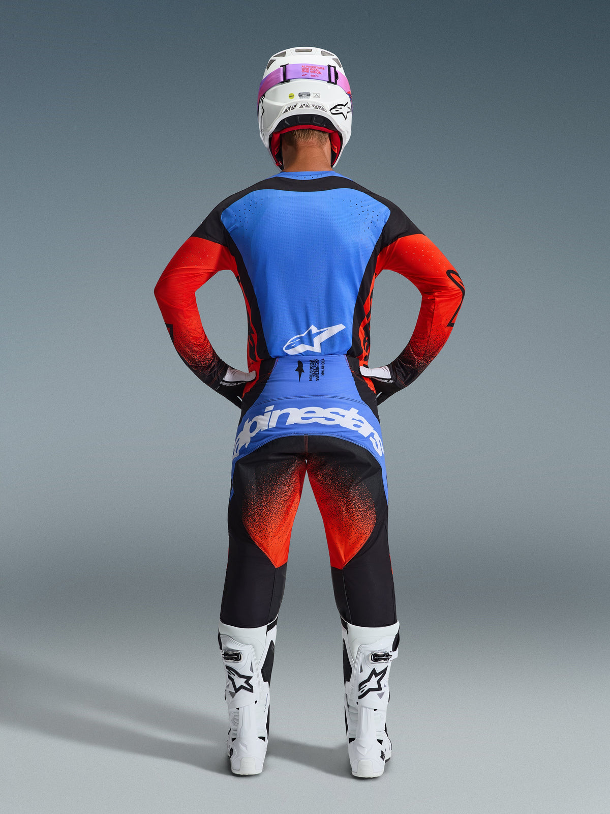 Vista posteriore di un pilota di motocross con il completo Alpinestars 2026 Techstar Knif Gear Set, maglia blu e rossa con dettagli neri, pantaloni neri con sfumatura rossa sulle cosce e logo blu, abbinato a stivali da corsa bianchi e casco bianco con maschera rosa.