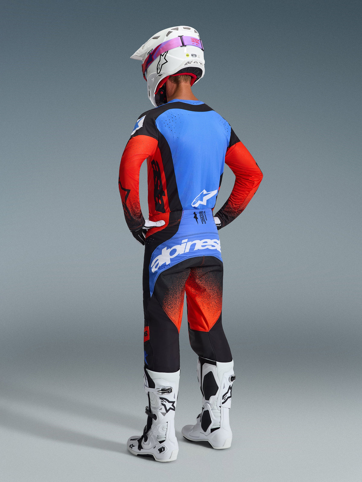 Vista posteriore di un pilota di motocross che indossa il completo Alpinestars 2026 Techstar Knif, caratterizzato da una maglia blu e rossa con accenti neri, pantaloni coordinati neri e rossi con un grande logo blu sulla seduta, casco bianco e stivali da motocross bianchi, kit da gara professionale per fuoristrada