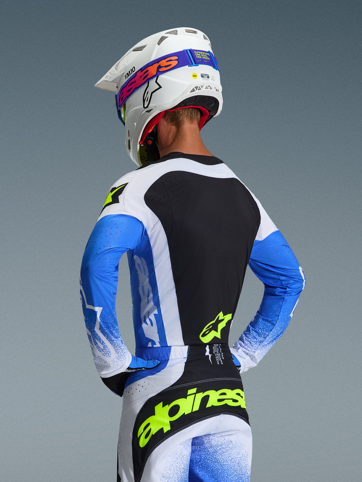 Vista posteriore di un pilota di motocross con maglia Alpinestars Techstar Knif 2026 in Blu Ucla, Nero e Giallo Fluo, abbinata a pantaloni coordinati e casco Supertech M10 bianco, abbigliamento tecnico con vestibilità racing per performance off-road.