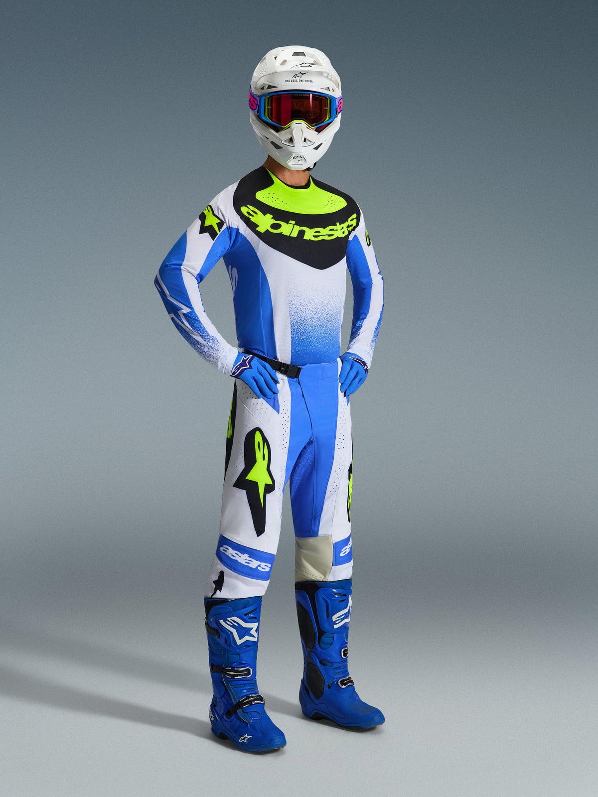 Pilota di motocross che indossa il completo Alpinestars Techstar Knif 2026 in Ucla Blue, nero e giallo fluo, composto da maglia tecnica, pantaloni coordinati con loghi evidenti, stivali blu, guanti blu e casco bianco con maschera, in piedi su sfondo grigio.