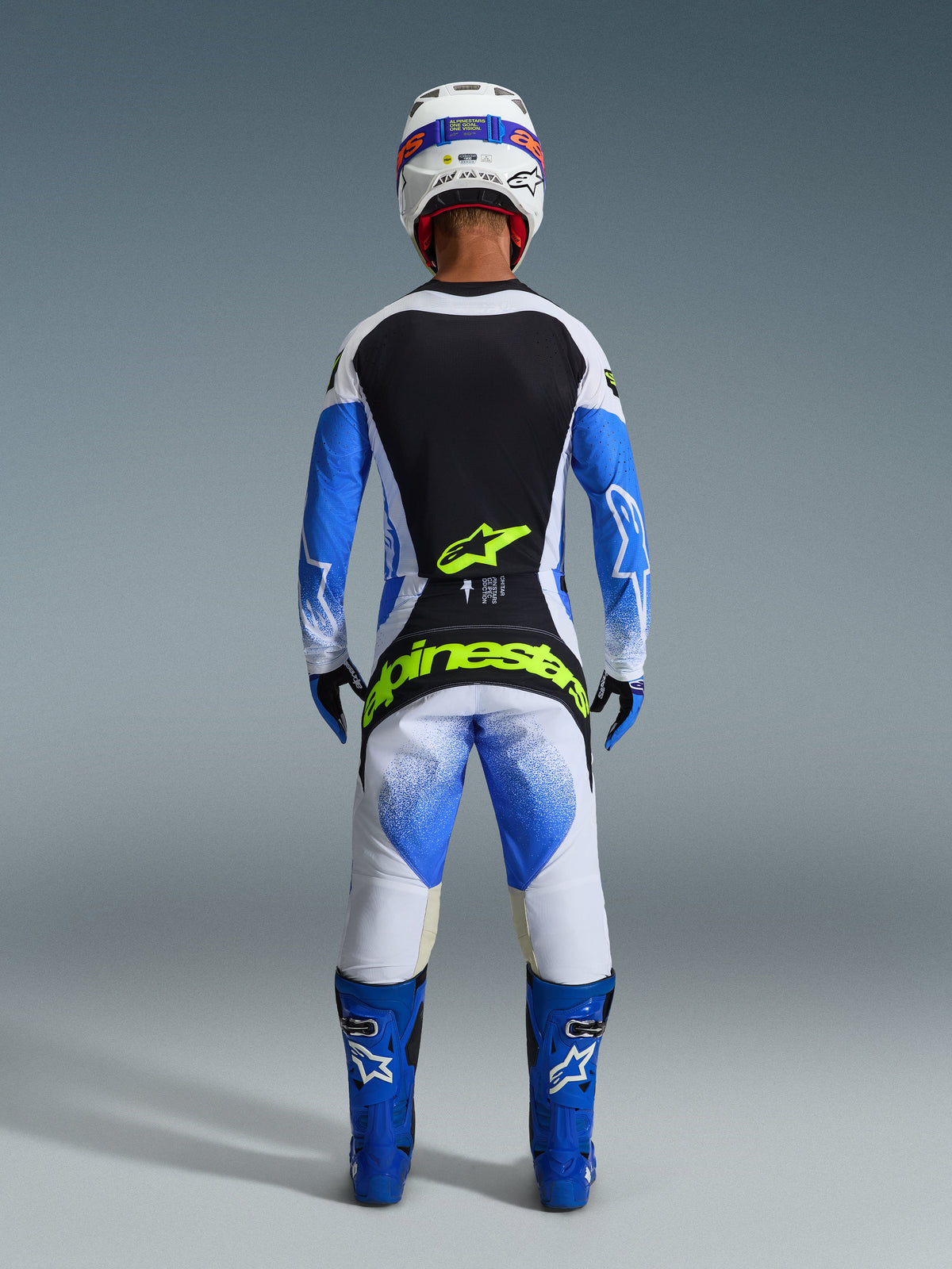 Alpinestars 2026 Techstar Knif Gear Set, vista posteriore di un pilota di motocross in blu Ucla, nero e bianco, con branding giallo fluo su maglia e pantaloni, abbinato a stivali da gara blu e casco bianco.