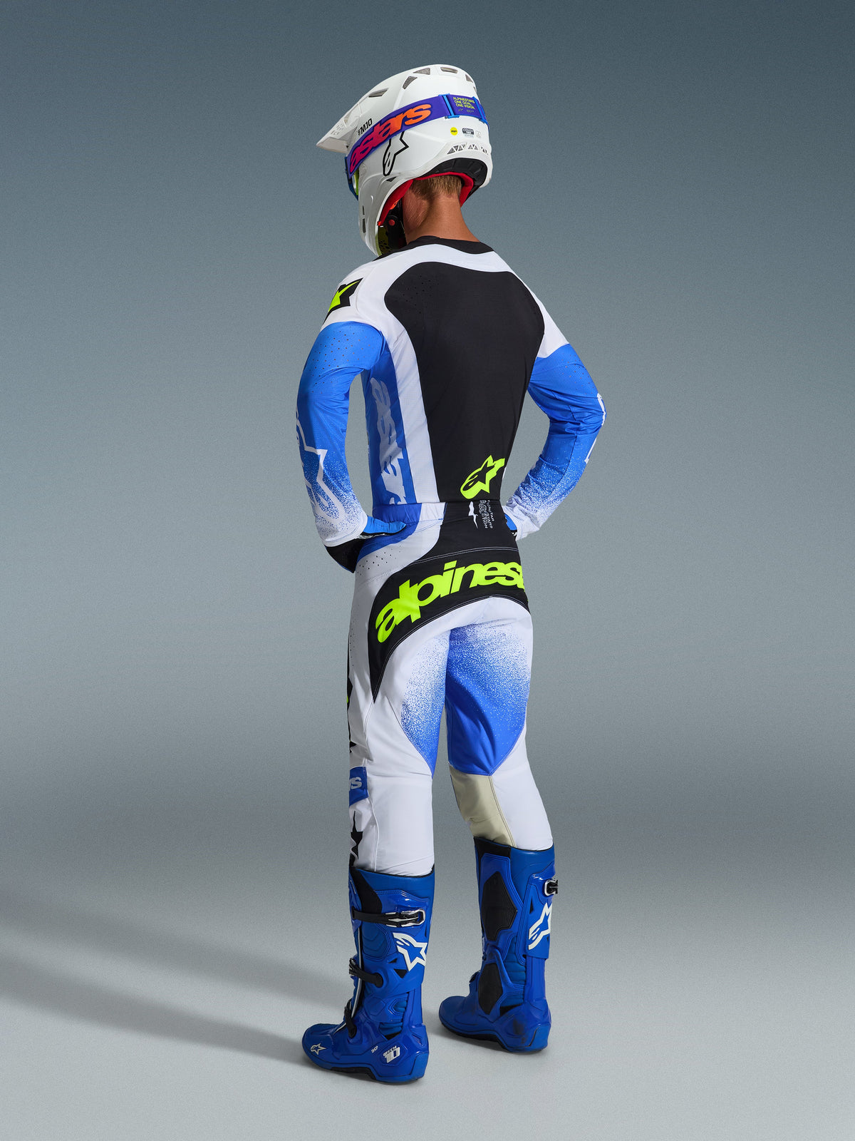 Pilota di motocross visto da dietro con il completo Alpinestars 2026 Techstar Knif in Blu Ucla e nero, maglia con pannello posteriore nero e logo giallo fluo, pantaloni coordinati blu e bianchi con branding giallo, stivali da motocross blu e casco bianco
