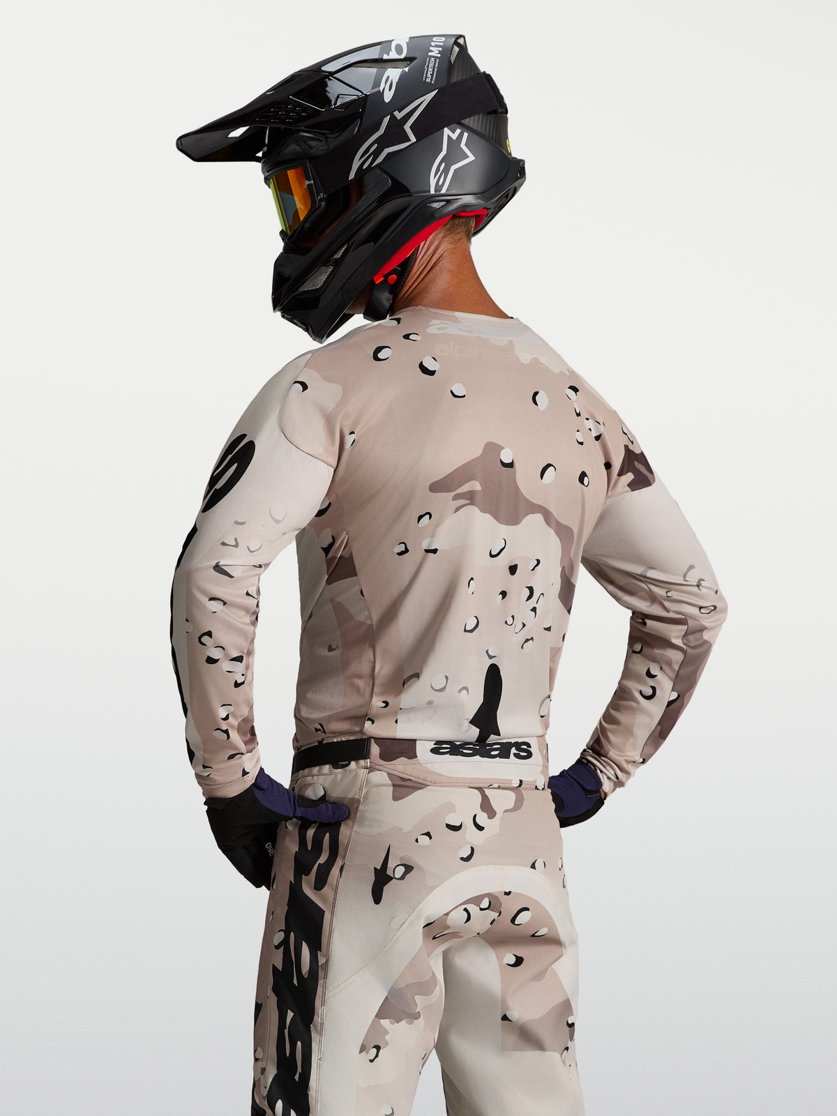 Alpinestars Maglia Racer Lone, maglia da motocross da uomo, sabbia e nero con motivo desert camo, vista posteriore su modello con casco, tessuto tecnico traspirante per sessioni off-road e enduro