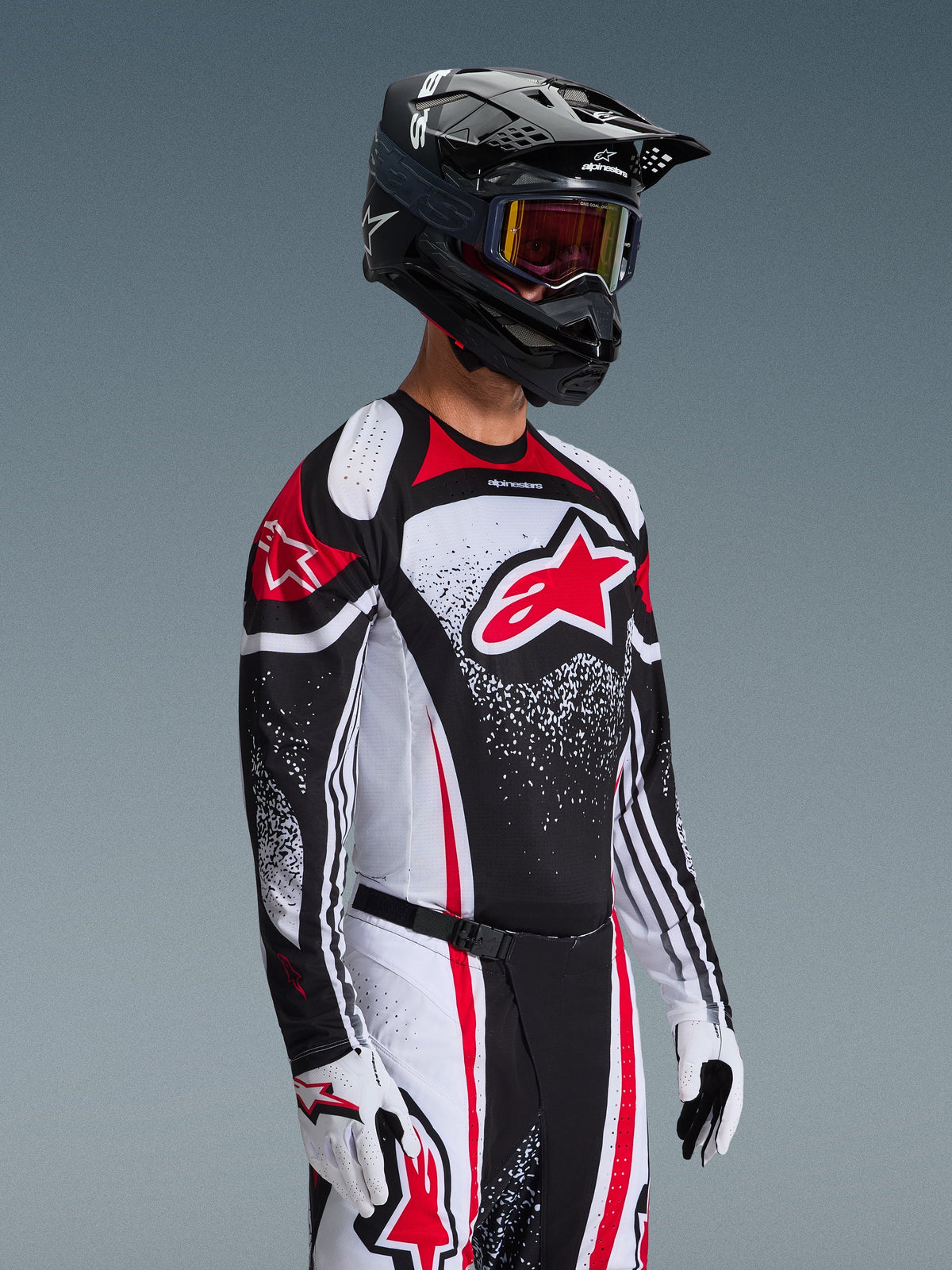 Pilota di motocross che indossa la maglia Alpinestars Techstar Nomur 2026 in nero bianco rosso, con un grande logo Astars rosso e grafiche puntinate, abbinata a pantaloni da gara coordinati, guanti bianchi e casco nero con maschera, completo coordinato per competizioni off-road