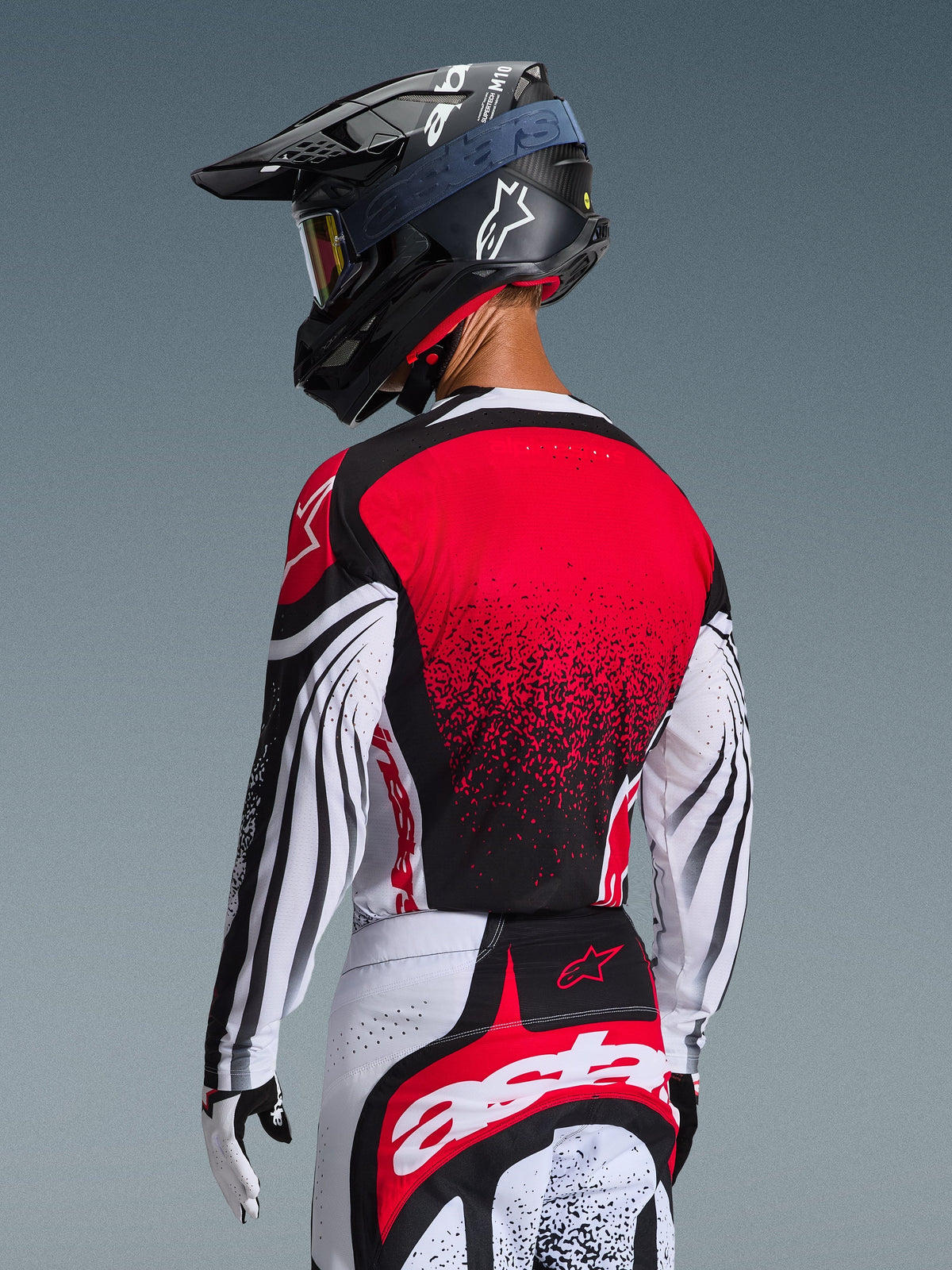 Pilota di motocross visto da dietro che indossa la maglia Techstar Nomur 2026, retro rosso con design sfumato nero e maniche bianche con accenti neri, abbinata a pantaloni rossi e bianchi coordinati, casco nero e guanti bianchi, abbigliamento professionale da corsa off-road