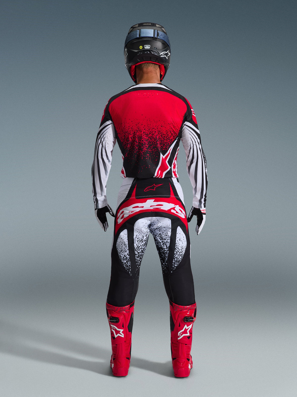 Pilota di motocross visto da dietro che indossa la maglia Alpinestars Techstar Nomur 2026, rosso, nero e bianco con motivo puntinato, pantaloni da gara coordinati, stivali da motocross rossi e casco nero, set completo di abbigliamento per competizioni off-road