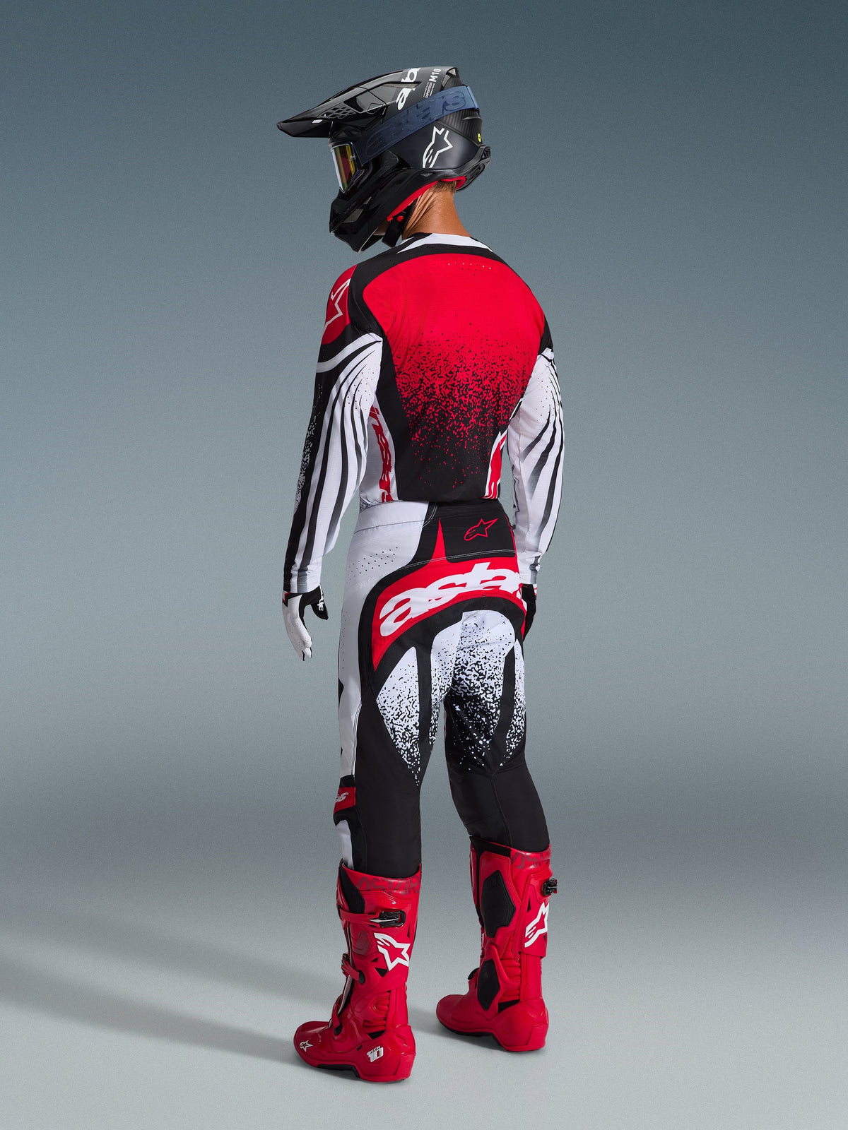 Vista posteriore di un pilota di motocross che indossa la maglia Alpinestars Techstar Nomur 2026 in Nero Bianco Rosso, caratterizzata da un retro con sfumatura rossa e motivi ondulati sulle maniche, abbinata a pantaloni da motocross coordinati, stivali rossi e un casco nero per un kit da gara off-road coordinato