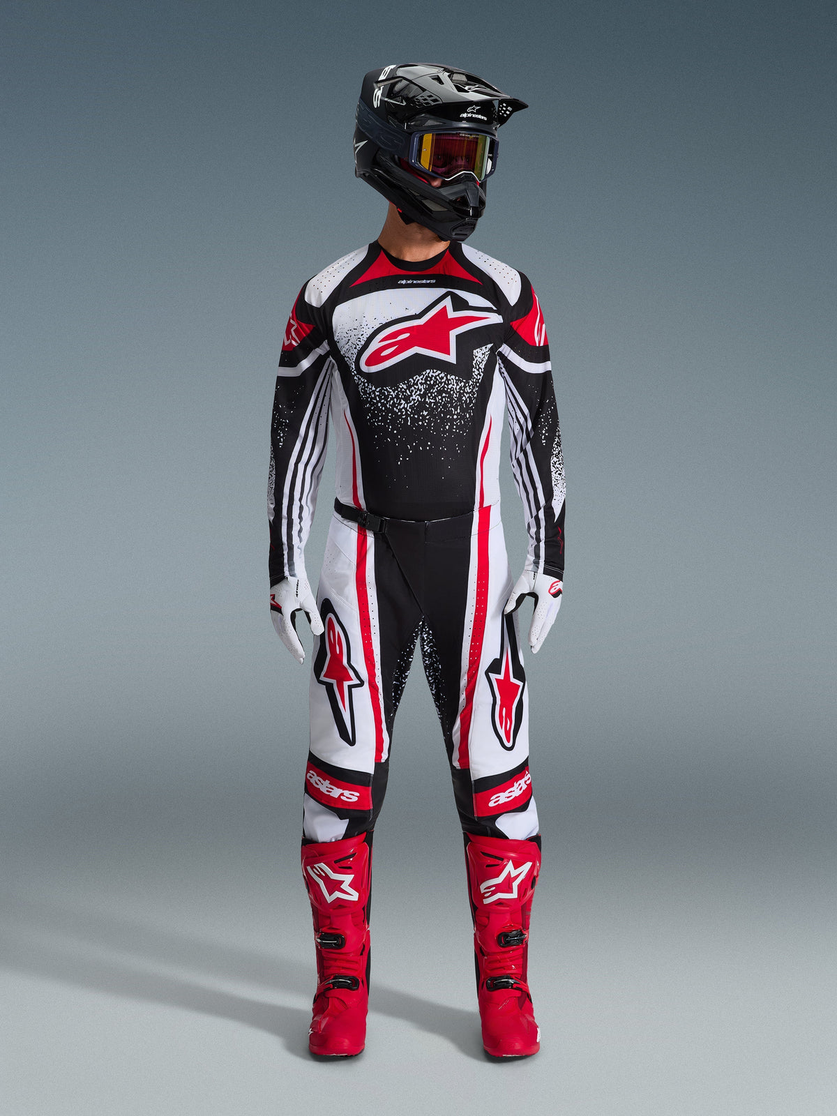 Pilota di motocross con abbigliamento completo da gara Alpinestars, maglia Techstar Nomur 2026 in nero, bianco e rosso, pantaloni coordinati con accenti rossi, casco nero e stivali rosso acceso, caratterizzato da un grande logo Astars rosso sul petto con motivo sfumato puntinato, completo per competizioni fuoristrada.
