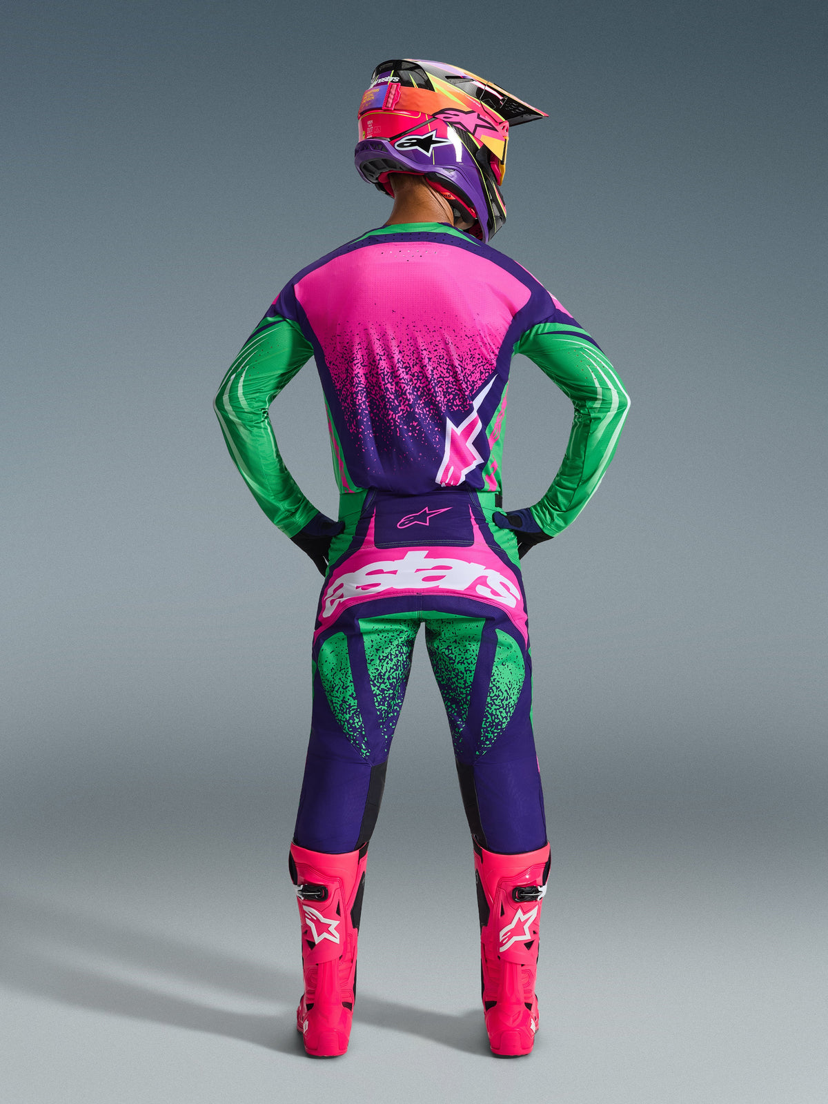 Set completo Alpinestars 2026 Techstar Nomur, pilota di motocross visto da dietro con maglia e pantaloni viola scuro, verde e rosa fluo, casco multicolore coordinato e stivali rosa fluo, outfit professionale da gara per prestazioni off-road