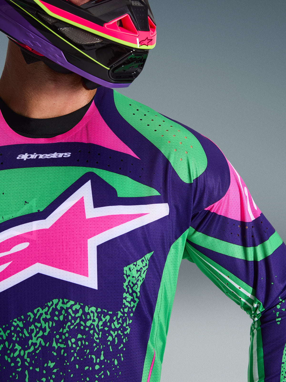 Primo piano della maglia Alpinestars 2026 Techstar Nomur, viola scuro con accenti verde e rosa fluo, con un grande logo a stella rosa sul petto e perforazioni tagliate al laser sulla spalla, indossata da un pilota con un casco da motocross coordinato viola e rosa.