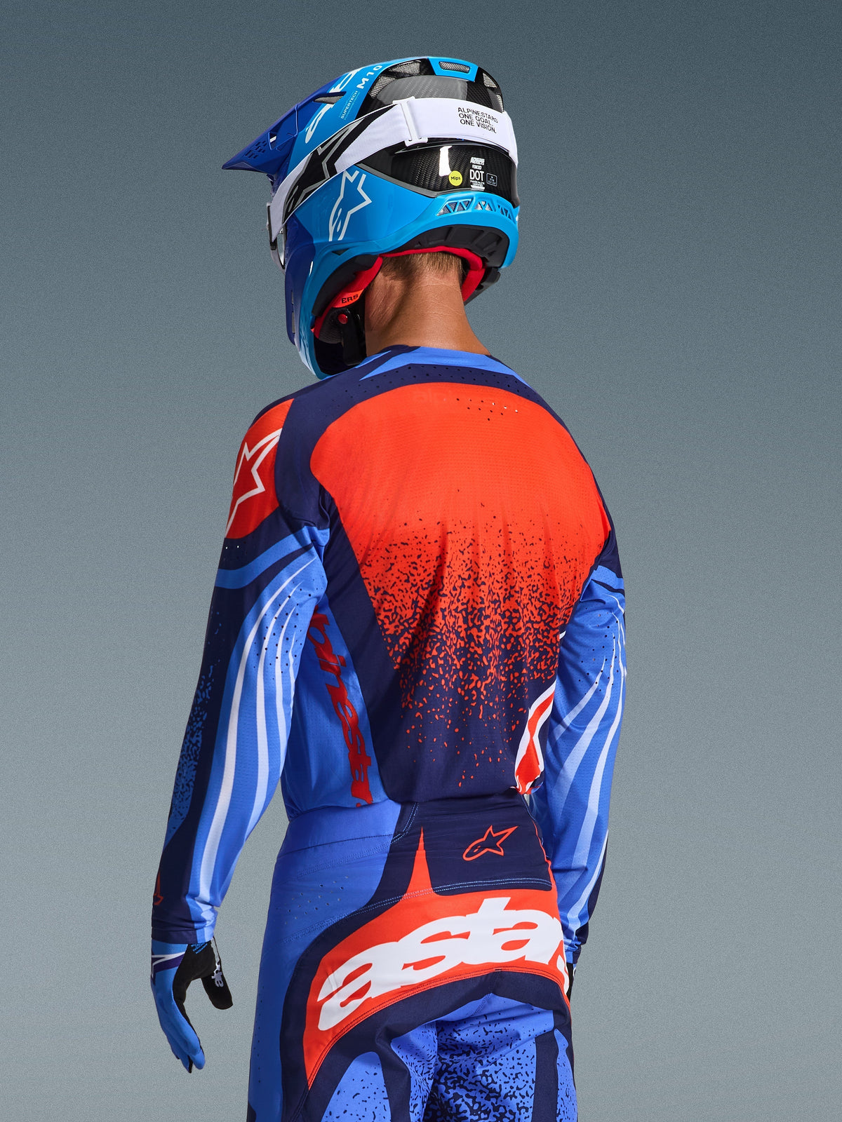 Pilota di motocross visto di spalle che indossa la maglia Alpinestars Techstar Nomur 2026 e i pantaloni coordinati, Dark Navy e UCLA Blue con vivaci accenti arancioni, con una grande grafica a stella e ventilazione tagliata al laser sulla maglia, abbinati a un casco blu e bianco per un kit da corsa off-road completo