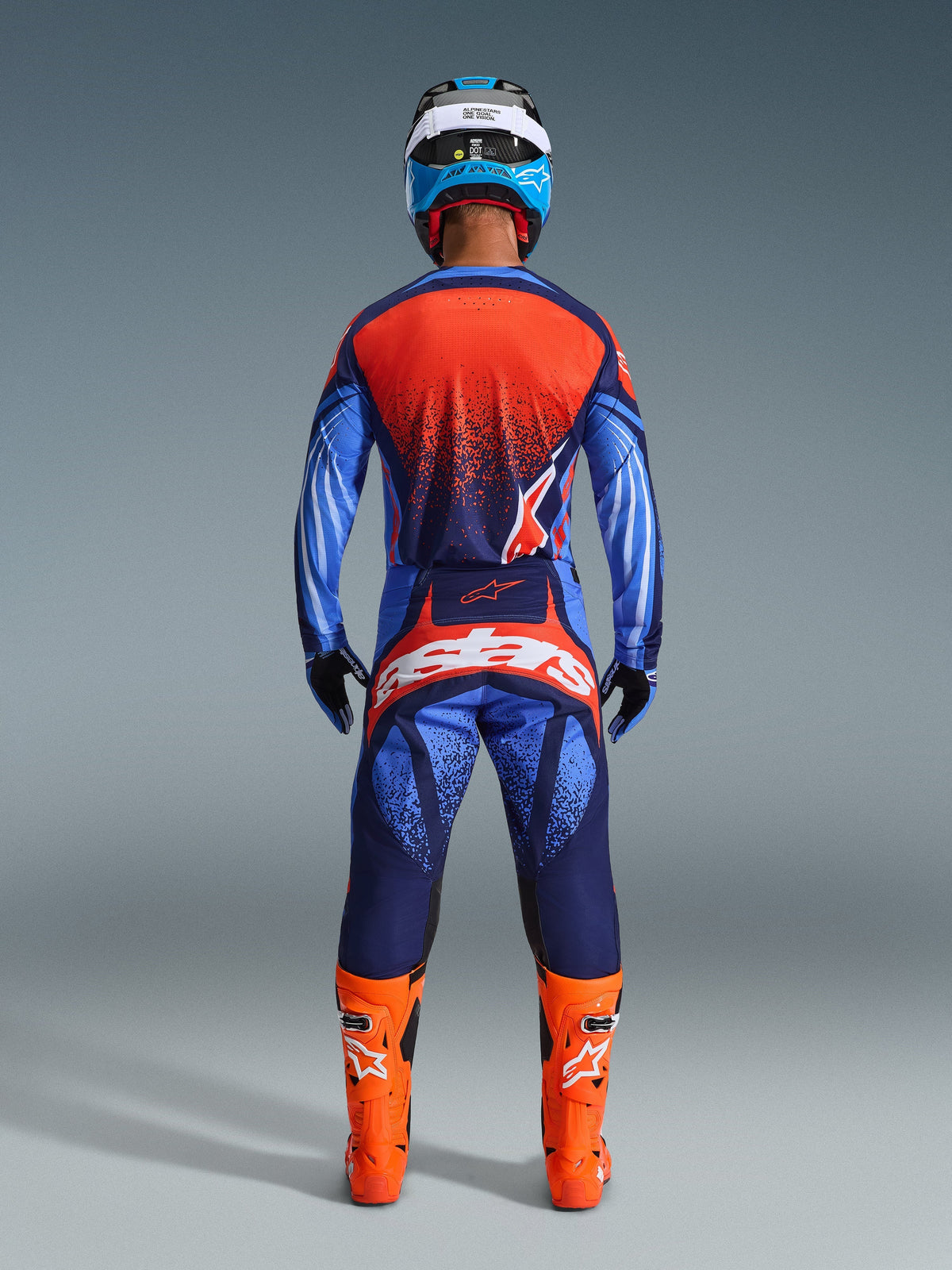 Pilota di motocross visto da dietro con il completo Alpinestars Techstar Nomur 2026 in blu scuro, arancione e blu UCLA, maglia con schiena arancione punteggiata, pantaloni coordinati con vistoso marchio bianco sulla seduta, stivali arancione acceso e casco blu e bianco, kit da corsa off-road coordinato.