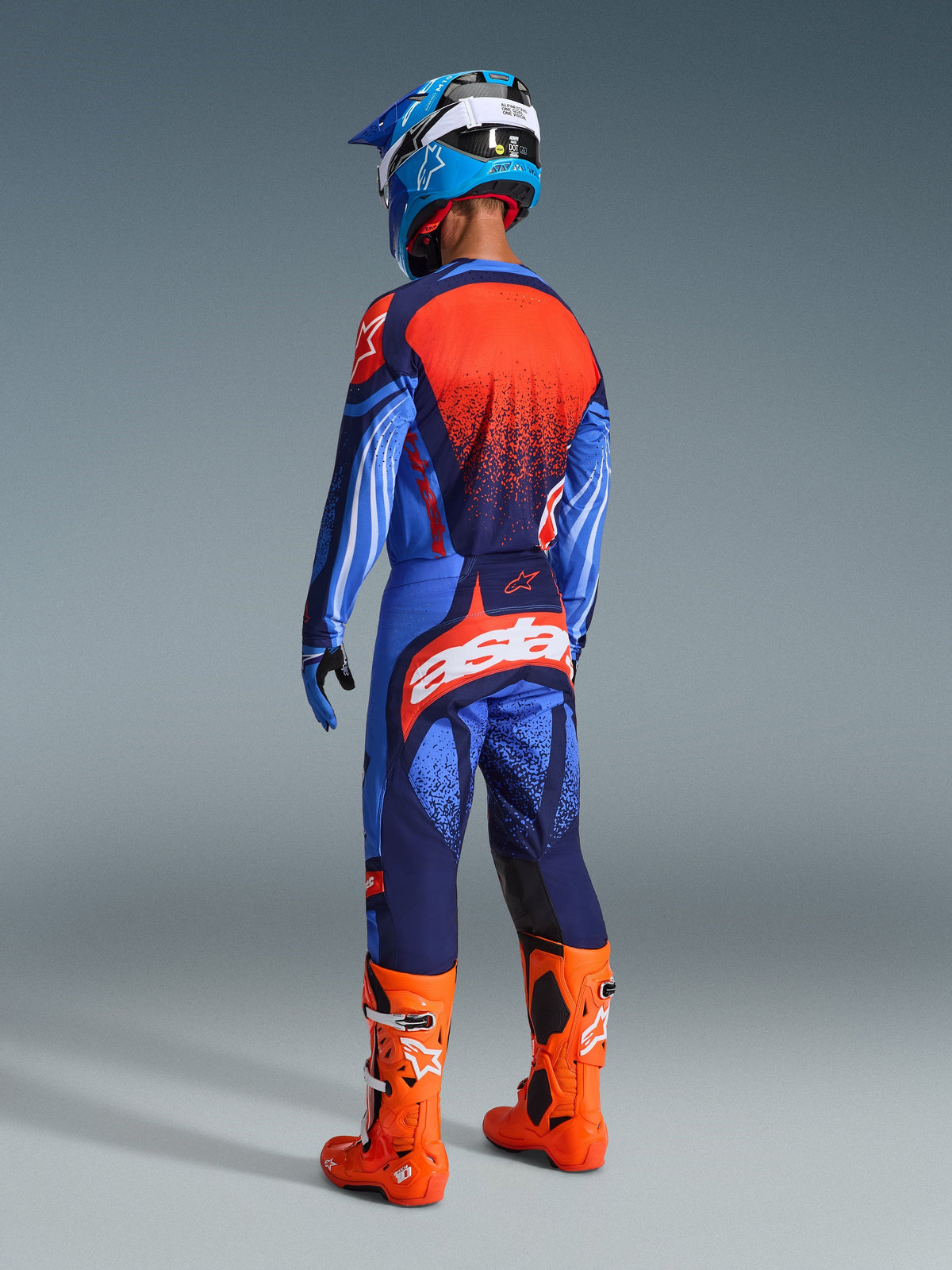 Pilota di motocross visto da dietro con completo Alpinestars Techstar Nomur 2026, maglia Dark Navy e blu UCLA con pannello arancione, pantaloni coordinati con logo 'astars' arancione, stivali da motocross arancione acceso e casco blu e bianco, abbigliamento da gara coordinato