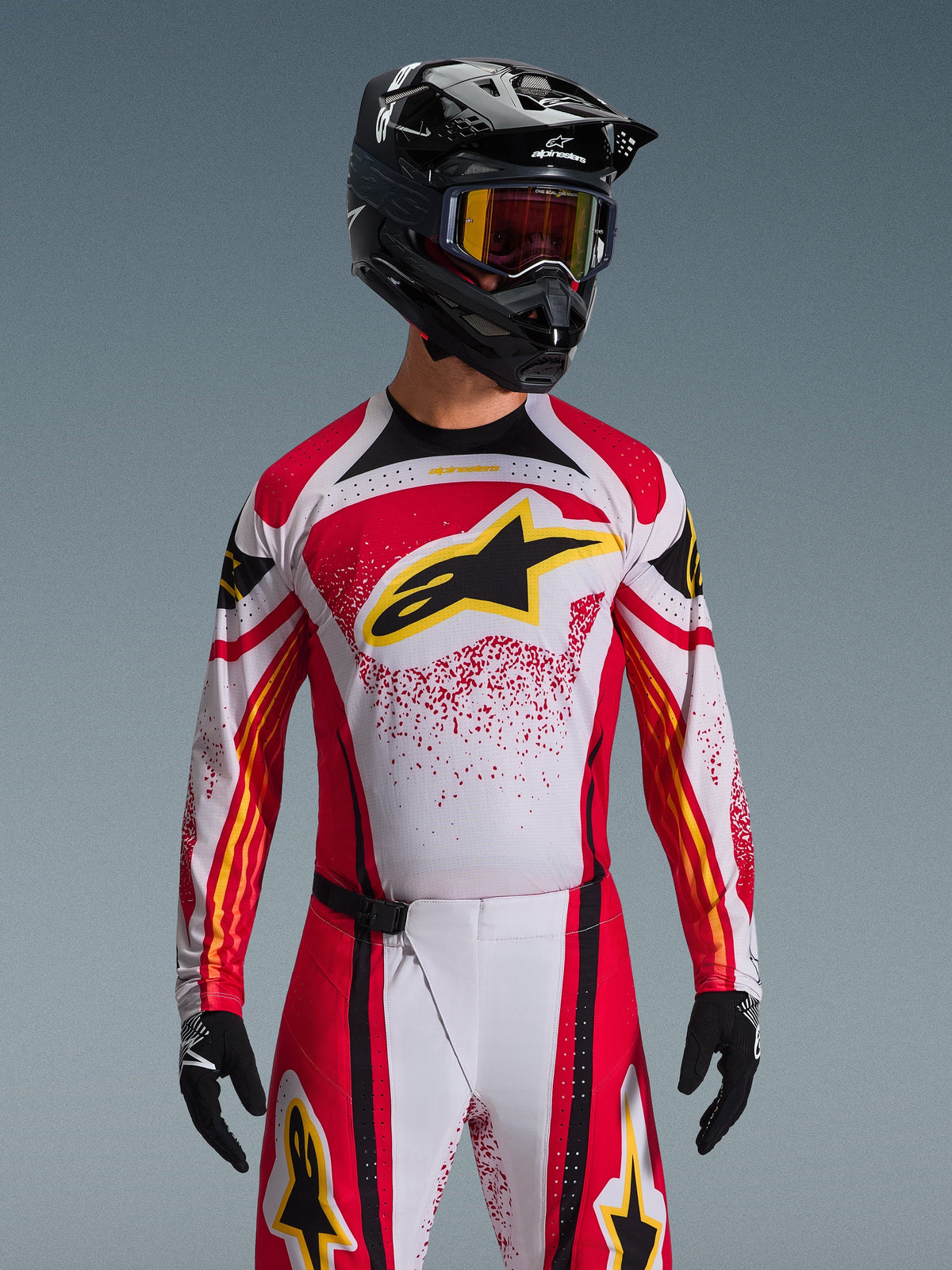 Alpinestars Techstar Nomur Jersey 2026, maglia da motocross, rosso, bianco e nero, con un grande logo A-star nero sul petto e accenti gialli, design con effetto splatter rosso, indossata con casco nero e attrezzatura off-road coordinata