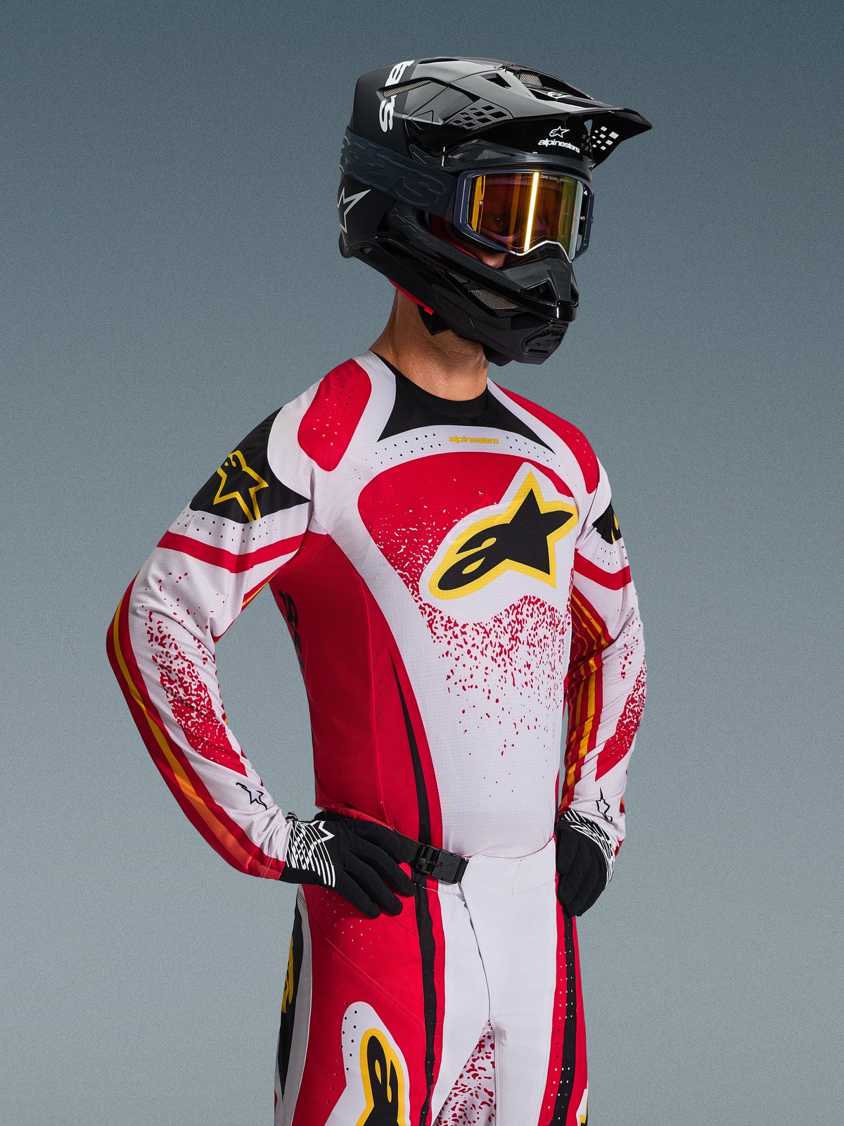 Pilota di motocross che indossa la maglia Alpinestars 2026 Techstar Nomur, rossa, bianca e nera con accenti gialli, abbinata a pantaloni coordinati, casco nero con maschera dorata e guanti neri, vestibilità atletica da gara per competizioni off-road professionali