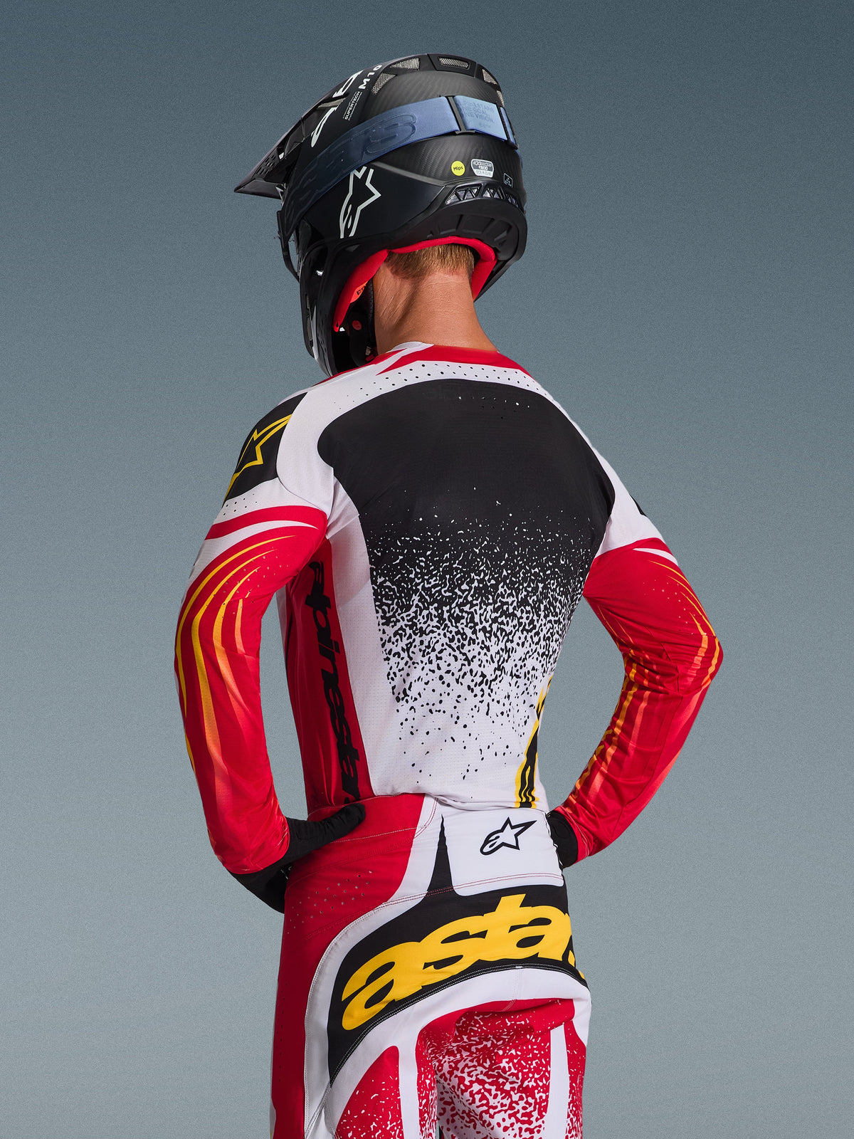Alpinestars 2026 Techstar Nomur Gear Set, maglia e pantaloni da motocross, rosso, bianco e nero con accenti gialli, vista posteriore del pilota con casco nero, abbigliamento racing con grafiche splatter e loghi evidenti