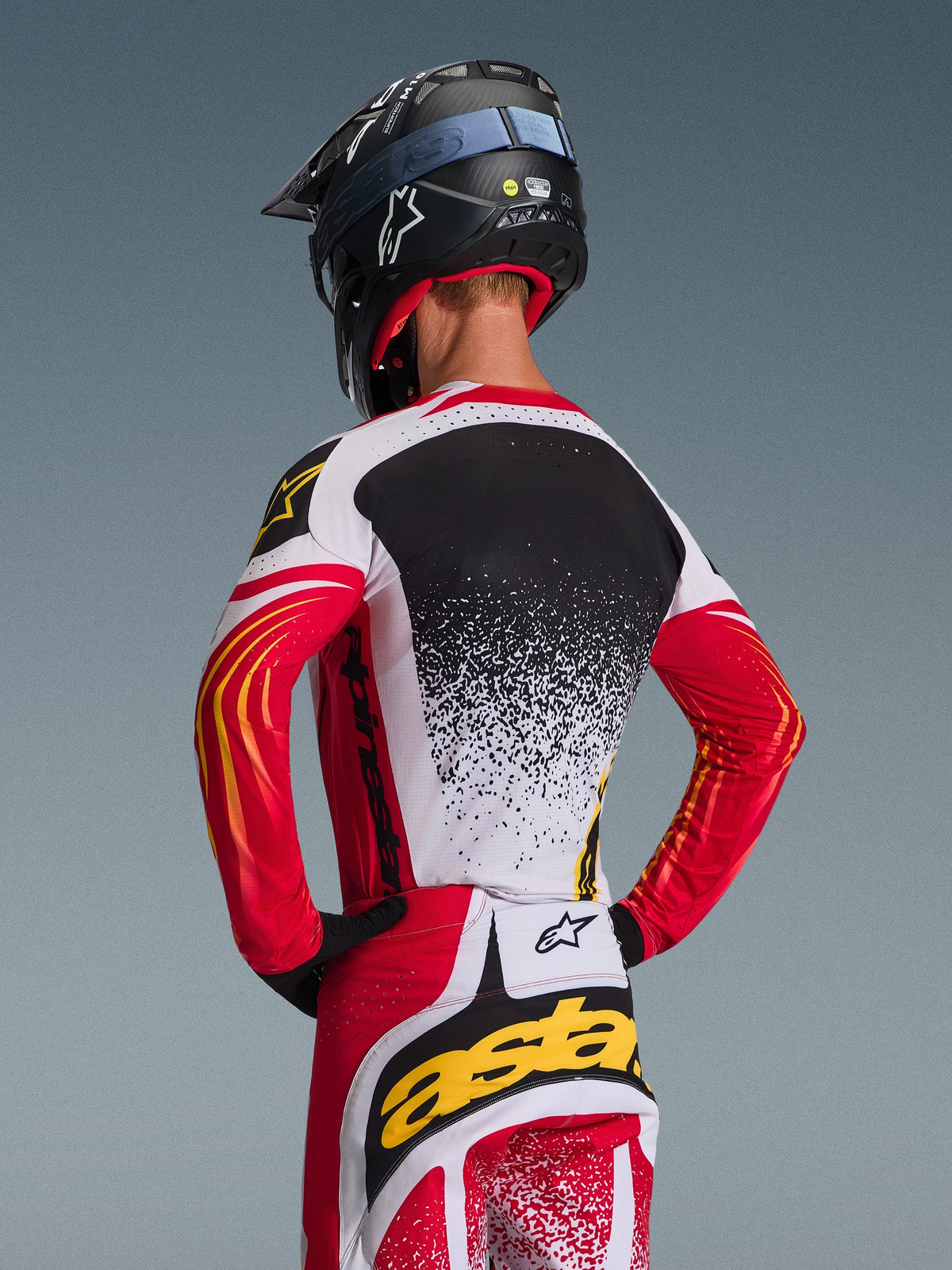 Pilota di motocross visto di spalle, maglia Alpinestars Techstar Nomur 2026 e pantaloni coordinati, rosso, bianco e nero con accenti gialli, design con sfumatura puntinata sulla schiena, casco Alpinestars nero e guanti neri, equipaggiamento professionale coordinato per gare fuoristrada