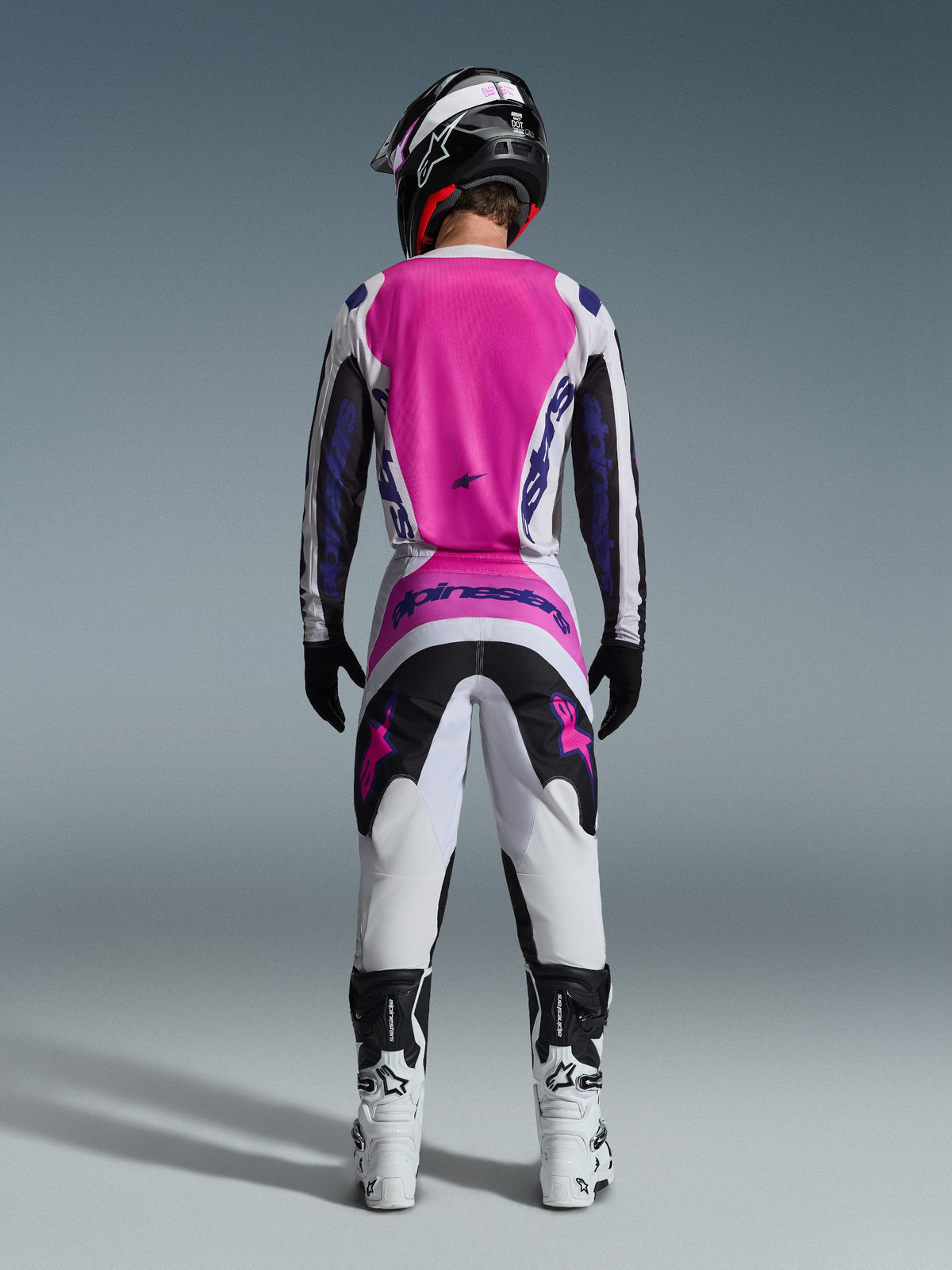 Pilota di motocross visto da dietro che indossa il set Alpinestars 2026 Fluid Grid, con maglia rosa acceso e bianca con maniche nere, pantaloni bianchi coordinati con pannelli neri e rosa, logo Alpinestars viola, stivali da gara bianchi e neri e casco abbinato, progettato per prestazioni off-road.