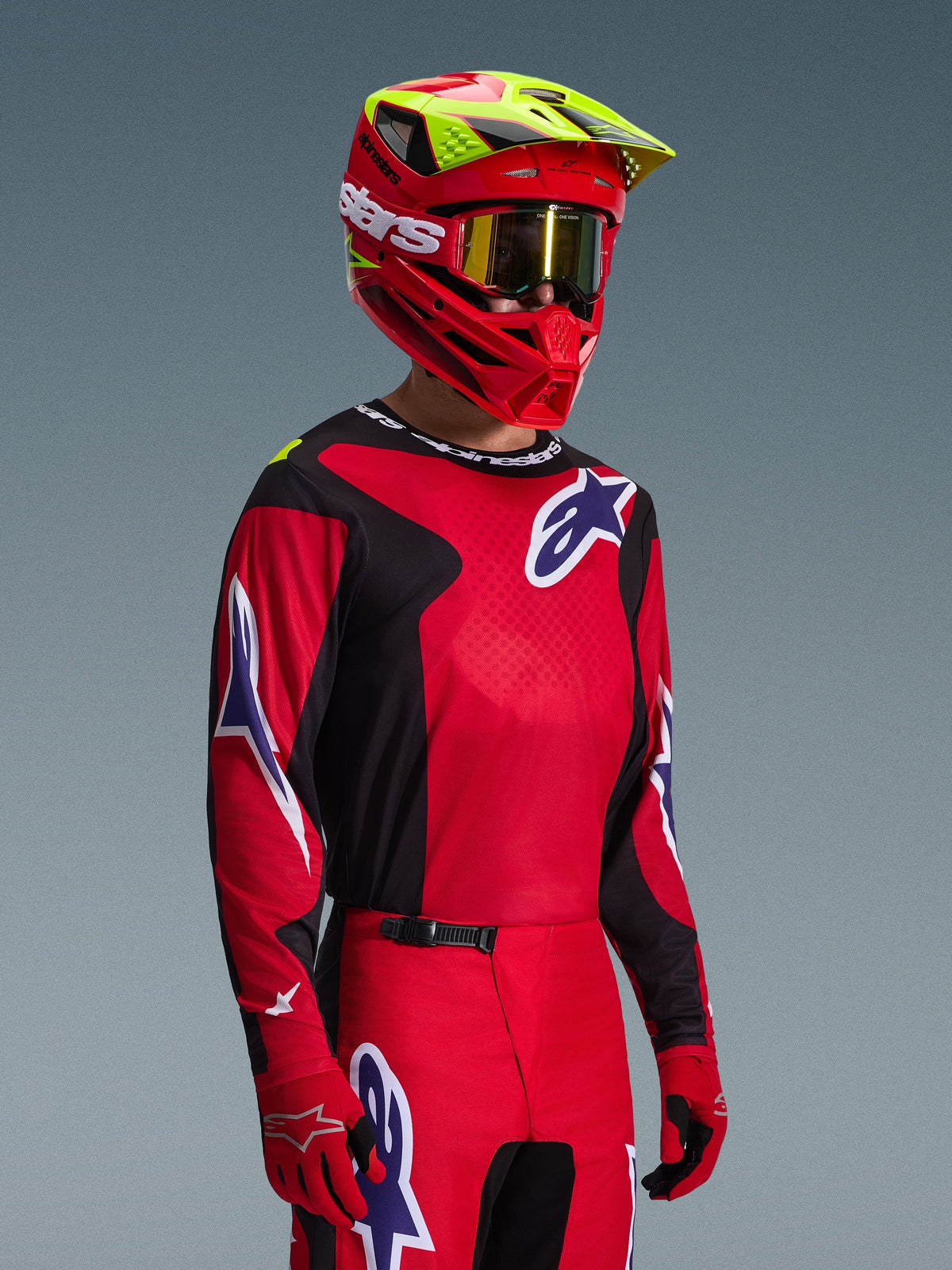 Pilota di motocross con maglia Alpinestars Fluid Grid 2026 in rosso, nero e viola, coordinata con pantaloni da motocross abbinati, casco rosso con accenti giallo neon, maschera dorata e guanti rossi, completo da gara off-road.
