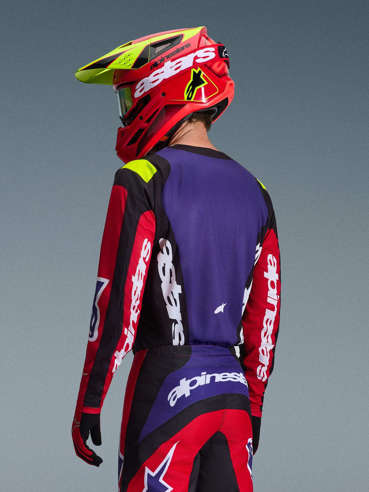 Pilota di motocross con maglia Alpinestars 2026 Fluid Grid, rosso, nero e viola, vista posteriore con pannello viola e maniche rosse con logo bianco, pantaloni coordinati e casco rosso e giallo neon, completo per corse fuoristrada