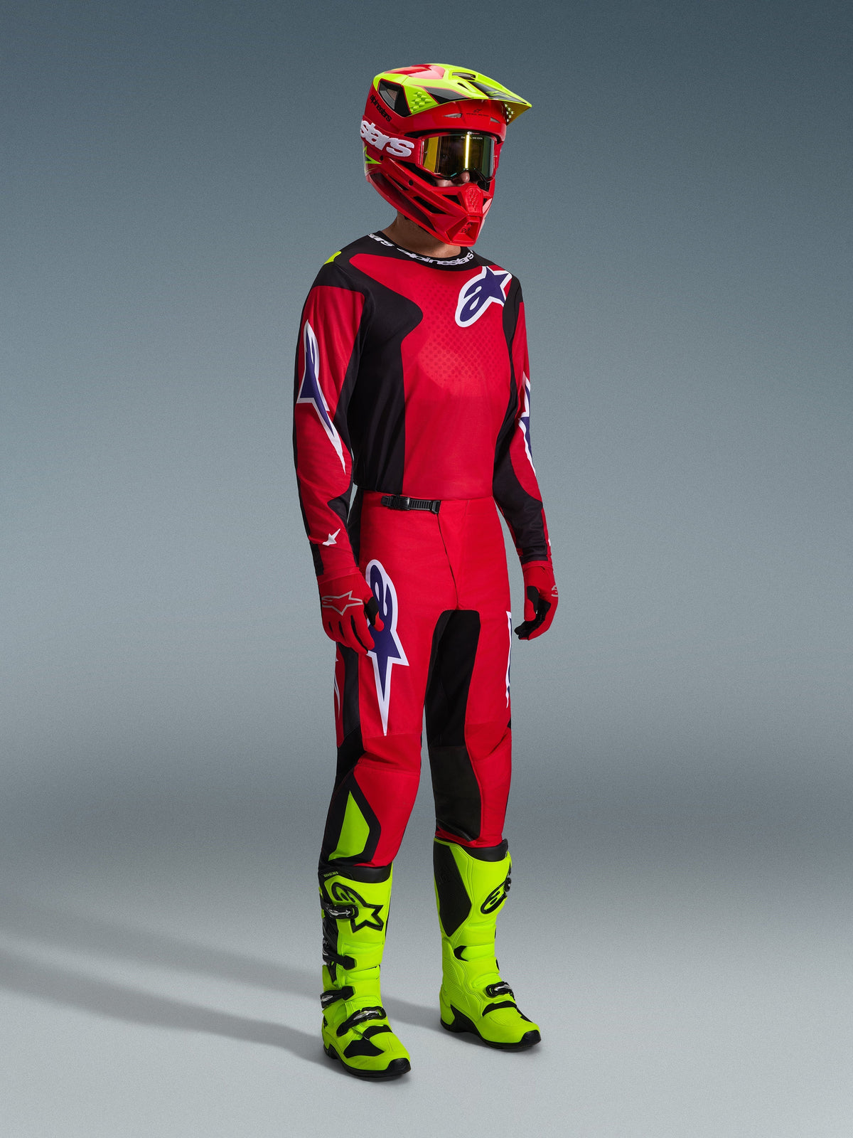 Pilota di motocross con abbigliamento completo da gara, maglia Fluid Grid 2026 in rosso, nero e viola, pantaloni coordinati con dettagli del logo viola, casco rosso e giallo neon con occhiali dorati e stivali giallo neon, outfit coordinato per competizioni fuoristrada