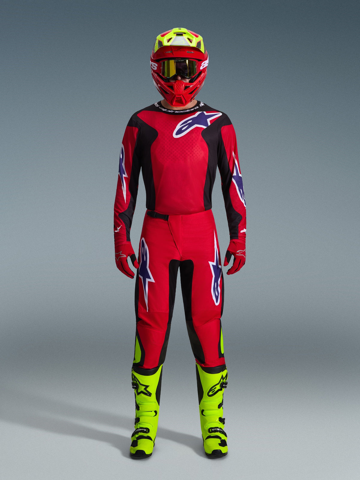 Pilota di motocross con Maglia Fluid Grid 2026 in rosso, nero e viola, pantaloni rossi coordinati con loghi viola, casco rosso con accenti giallo neon e stivali giallo neon, kit da gara completo per motocross off-road