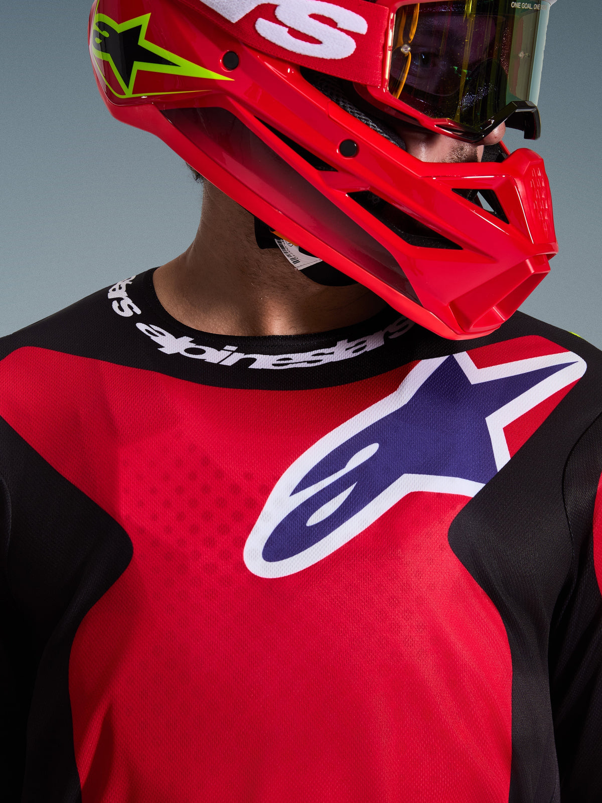 Primo piano di un pilota di motocross che indossa la maglia 2026 Fluid Grid in rosso, nero e viola, caratterizzata da un grande logo Alpinestars sul petto e colletto sportivo logato, abbinata a un casco rosso e maschera, che mette in risalto il tessuto tecnico e la vestibilità atletica.