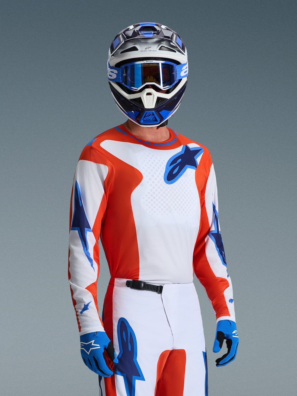 Alpinestars Maglia Fluid Grid 2026, maglia da motocross in Orange UCLA Blue, indossata da un pilota con pantaloni bianchi e arancioni coordinati, casco bianco e blu, maschera blu e guanti blu, completo da gara off-road