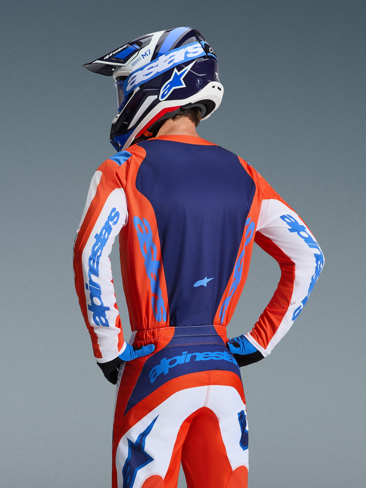 Maglia Alpinestars Fluid Grid 2026, maglia da motocross, arancione e blu UCLA, vista posteriore del pilota con pantaloni e casco coordinati, con pannello posteriore blu navy e maniche bianche con marchio azzurro, vestibilità atletica per corse fuoristrada