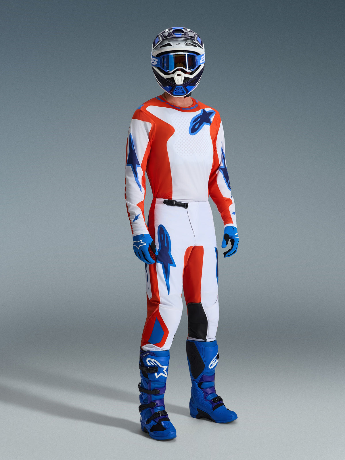 Pilota di motocross con abbigliamento da gara completo, maglia Fluid Grid 2026 in arancione e blu UCLA, pantaloni coordinati con pannelli bianchi e loghi blu, stivali da motocross blu e casco coordinato, scatto professionale in studio per gare off-road