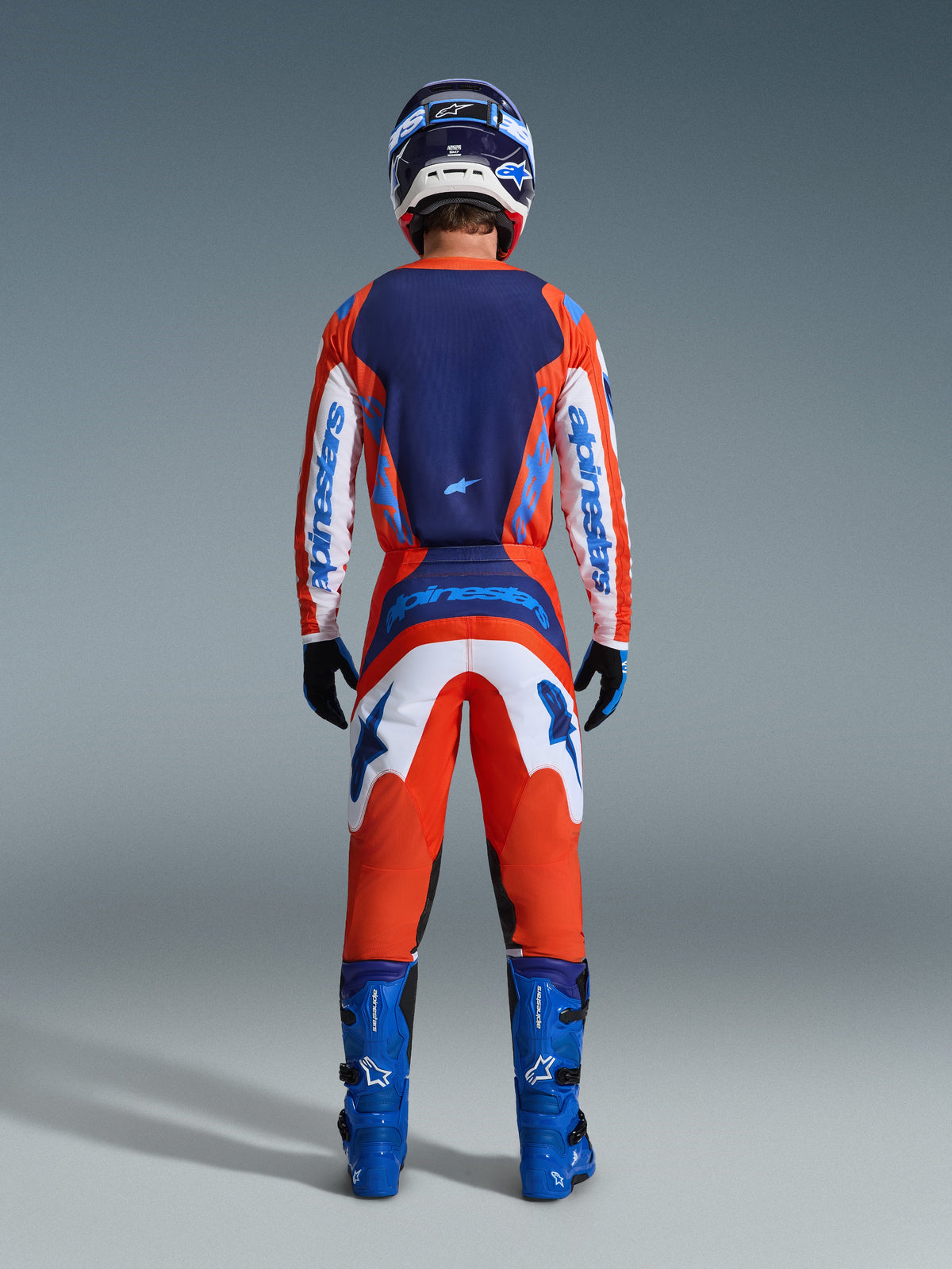 Alpinestars Maglia Fluid Grid 2026, maglia da motocross, arancione e blu UCLA, vista posteriore del pilota con set completo da gara, caratterizzata da un pannello posteriore blu scuro e maniche bianche con loghi blu, abbinata a pantaloni coordinati e stivali blu