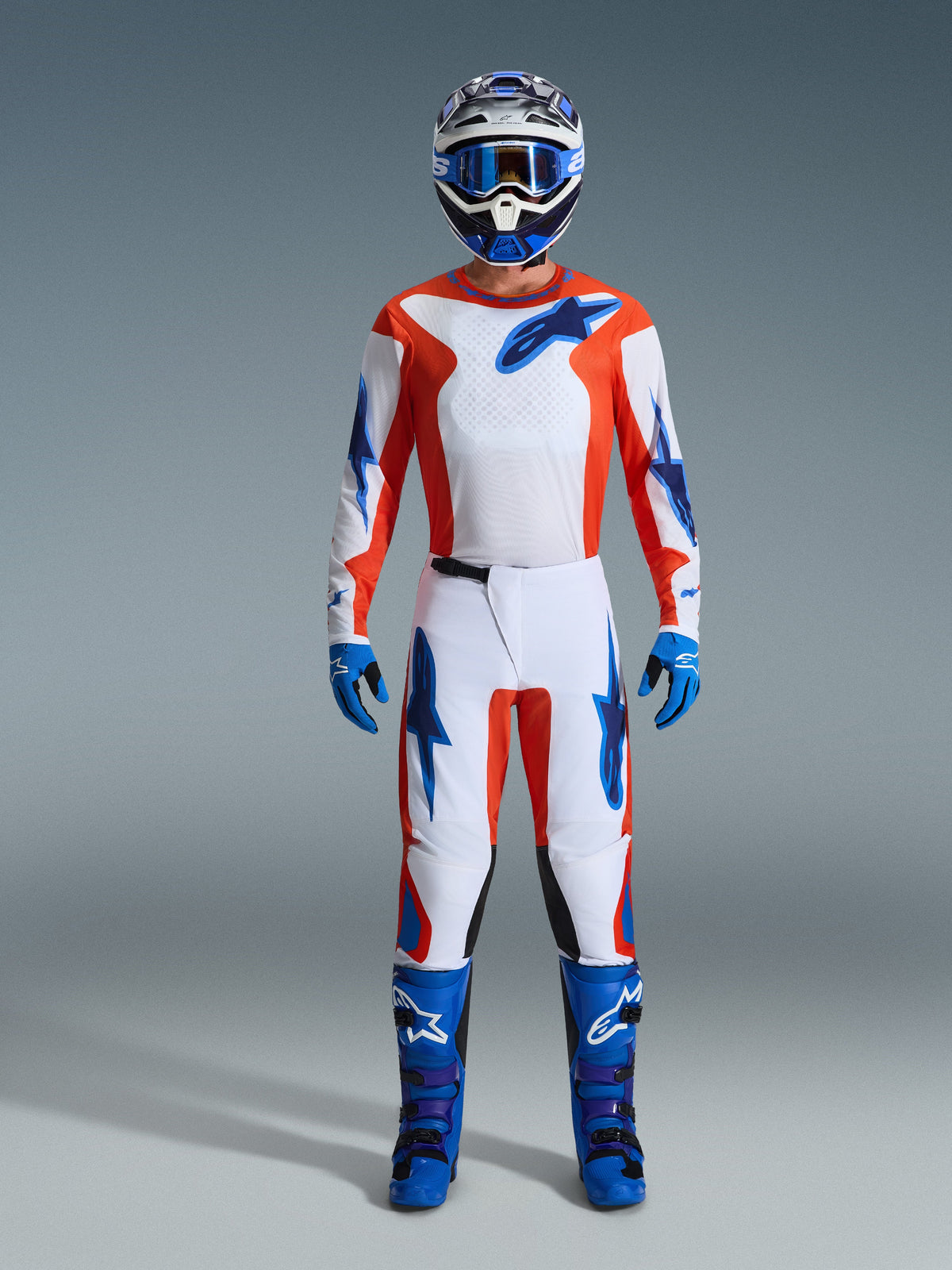 Pilota di motocross con equipaggiamento da gara completo, maglia Fluid Grid 2026 in arancione e blu UCLA, pantaloni bianchi coordinati con accenti arancioni e blu, casco blu e bianco con maschera blu, stivali da fuoristrada blu e viola, guanti blu, completo coordinato per corse di motocross