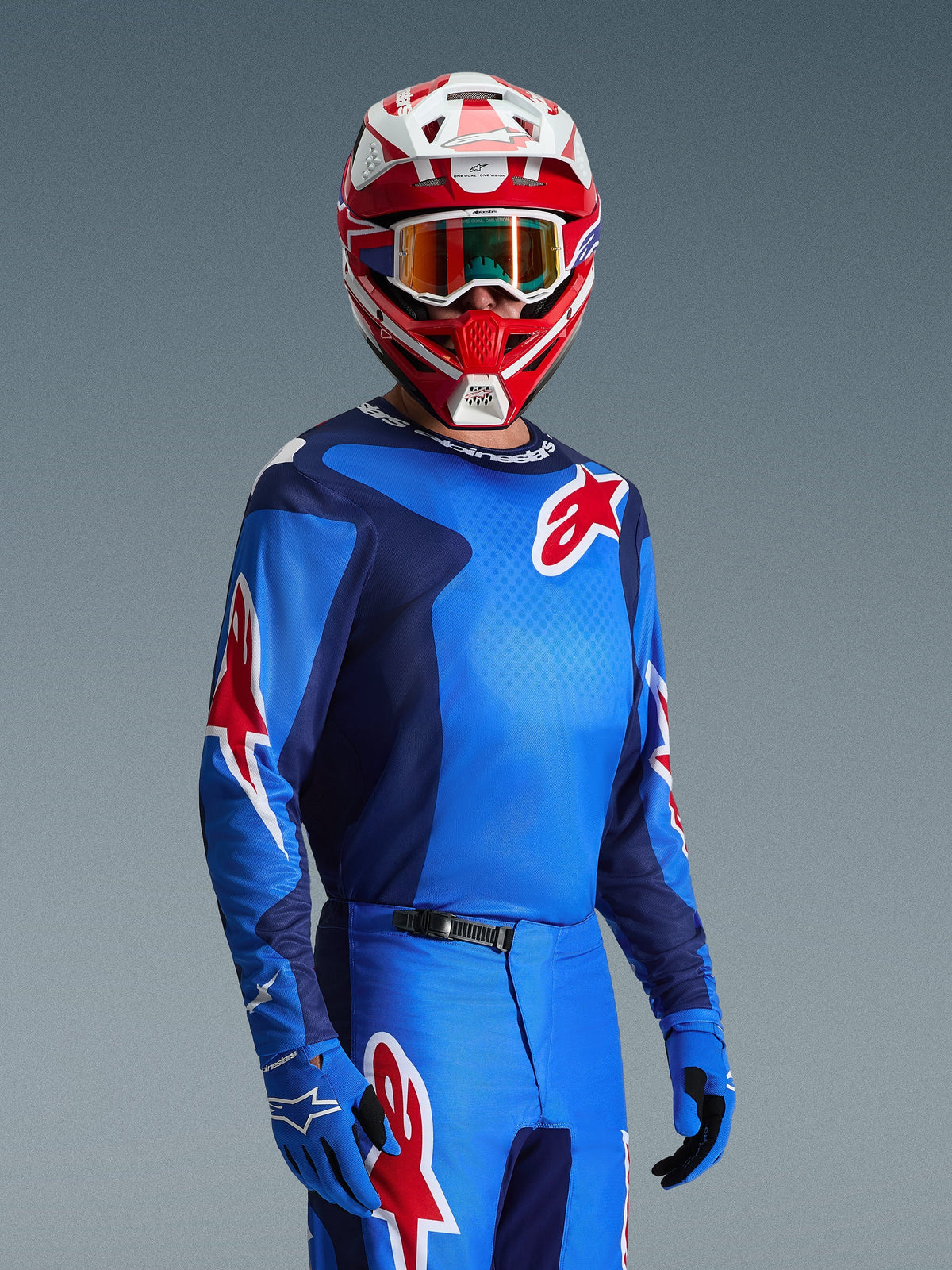 Pilota di motocross con maglia Alpinestars Fluid Grid 2026 in blu e rosso, con loghi a stella Alpinestars rossi, abbinata a pantaloni blu coordinati, guanti blu e un casco rosso e bianco con maschera
