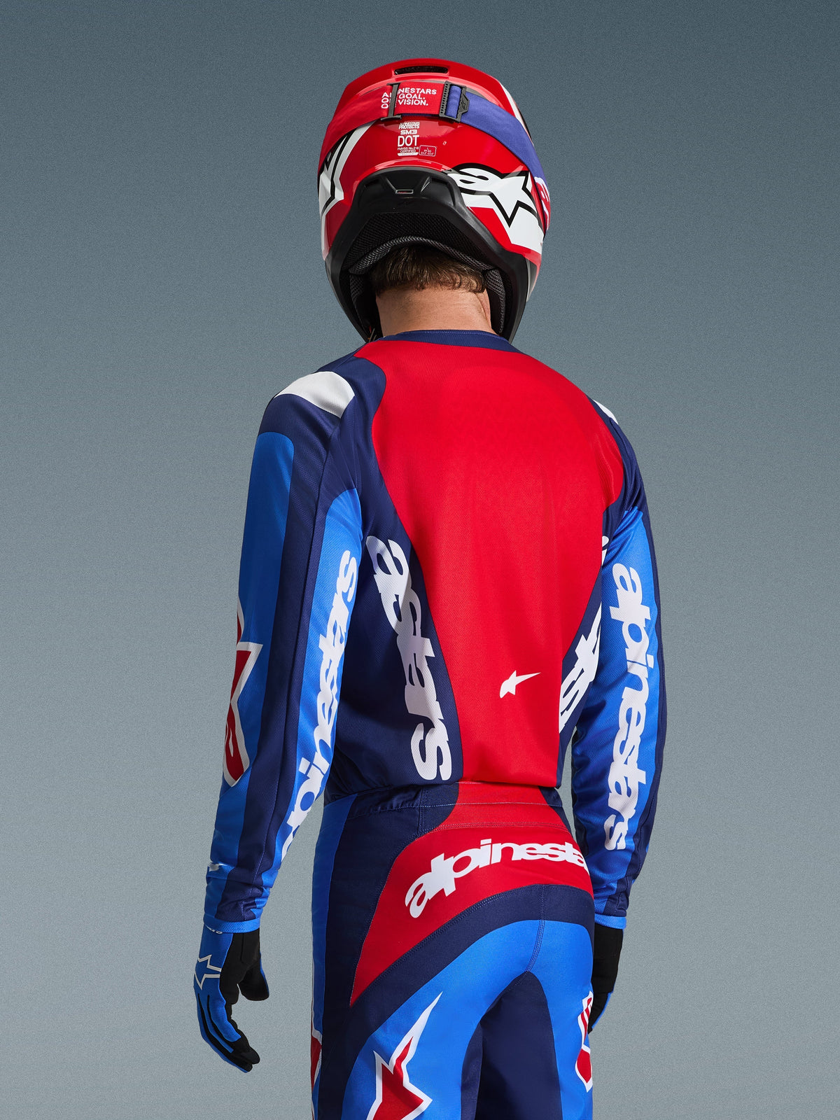 Vista posteriore di un pilota di motocross che indossa la maglia Alpinestars 2026 Fluid Grid in Blu e Rosso, caratterizzata da loghi bianchi su maniche e schiena, abbinata a pantaloni da motocross coordinati e un casco rosso per un look da gara off-road completo