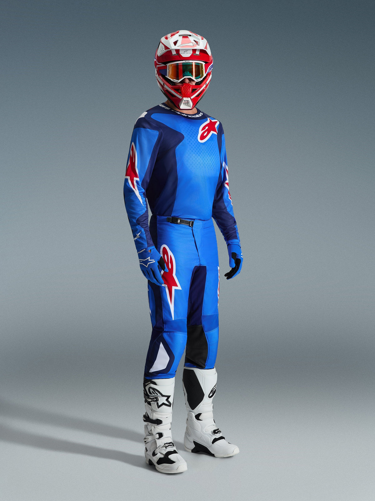 Pilota di motocross che indossa la maglia Alpinestars 2026 Fluid Grid in Blue Red, abbinata a pantaloni da motocross coordinati, stivali bianchi e neri e un casco rosso e bianco, set di abbigliamento professionale per prestazioni off-road.