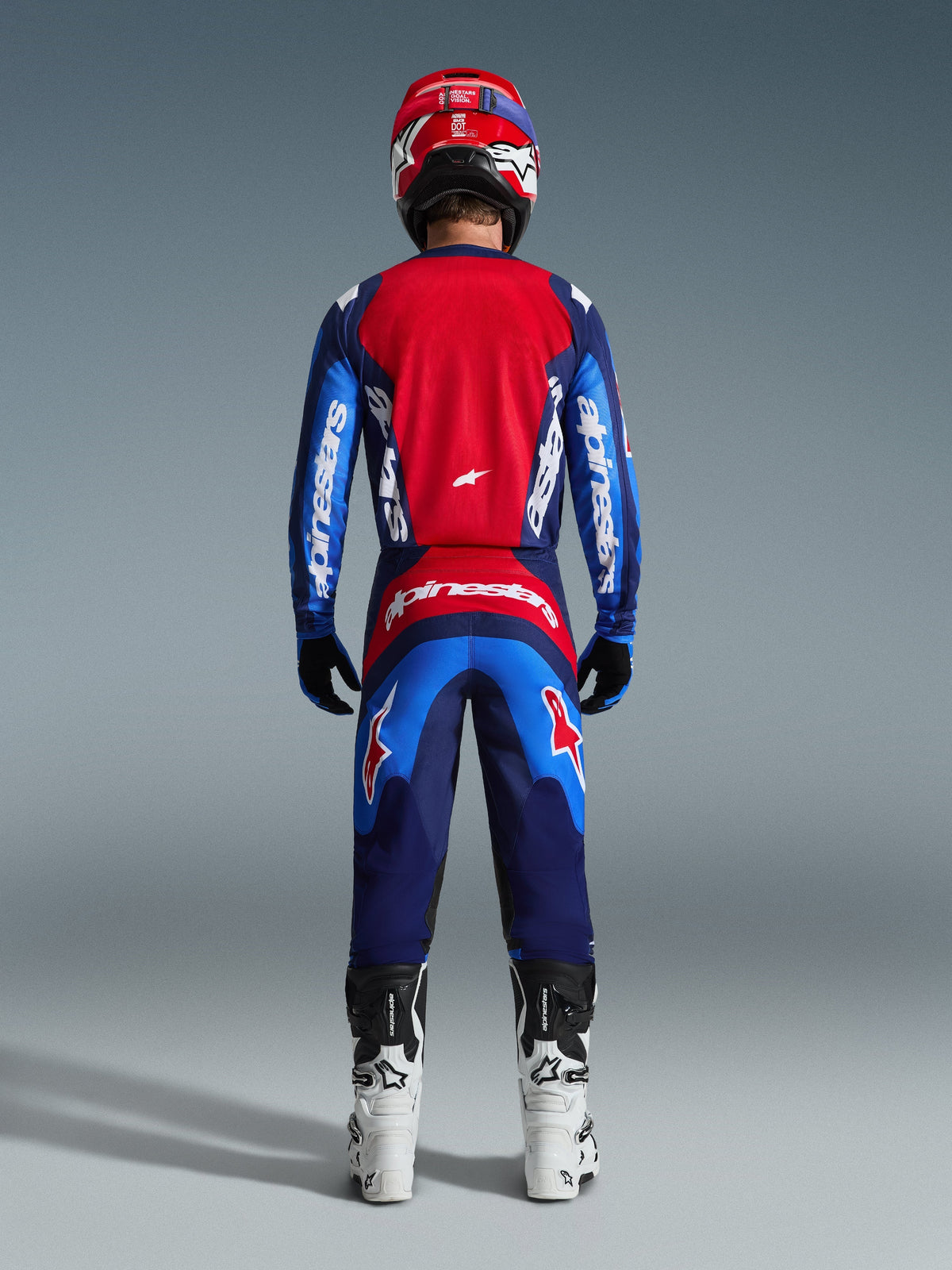 Vista posteriore di un pilota di motocross che indossa la Maglia Fluid Grid 2026 in blu e rosso, caratterizzata da un ampio pannello posteriore rosso e maniche blu con marchio Alpinestars bianco, abbinata a pantaloni coordinati, stivali bianchi e neri e un casco rosso per un completo da gara off-road.