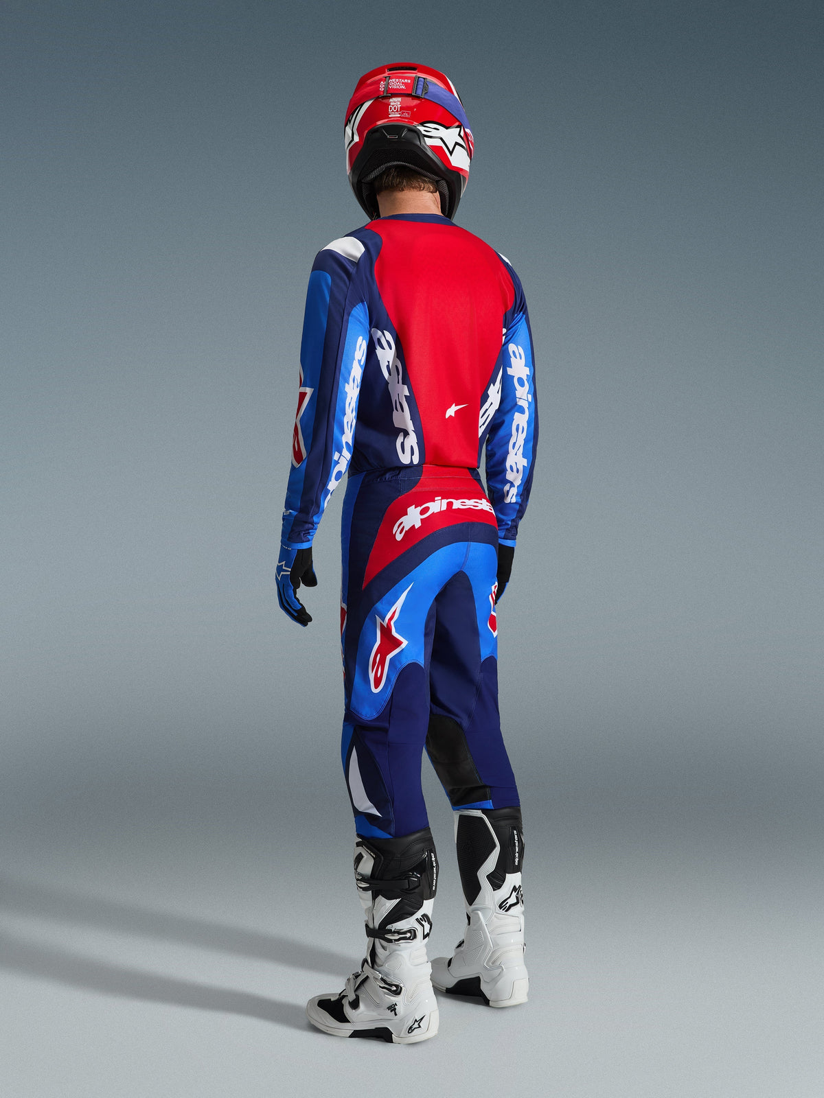 Pilota di motocross visto di spalle che indossa la maglia Alpinestars 2026 Fluid Grid in blu e rosso, pantaloni da motocross coordinati con branding bianco in evidenza, casco rosso e stivali bianchi e neri, set completo di equipaggiamento da gara off-road