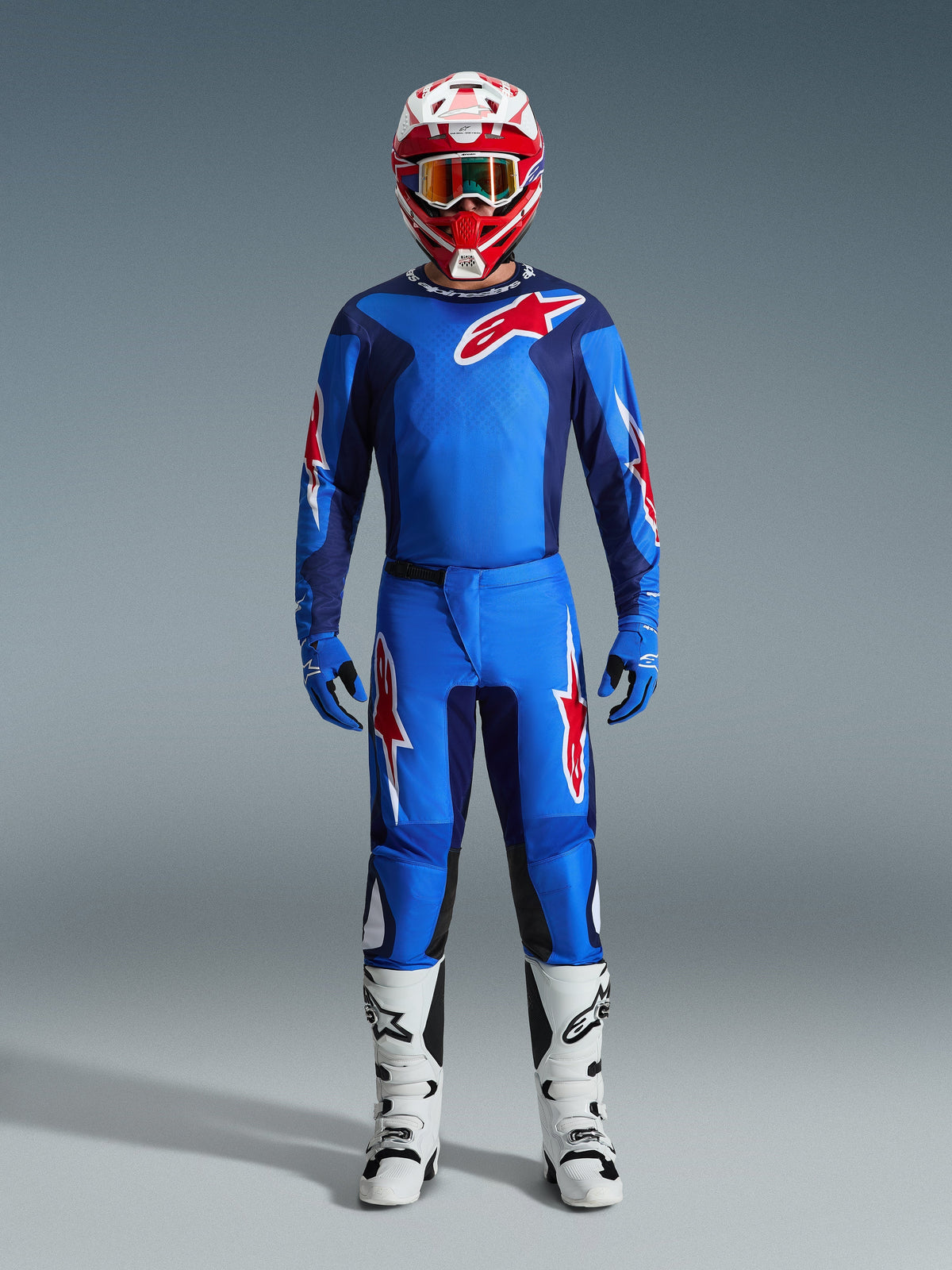 Pilota di motocross con maglia Alpinestars 2026 Fluid Grid in blu e rosso, pantaloni blu coordinati con loghi a stella bianchi e rossi, casco rosso e bianco con maschera a specchio, guanti blu e stivali da corsa bianchi e neri, set completo di abbigliamento fuoristrada.
