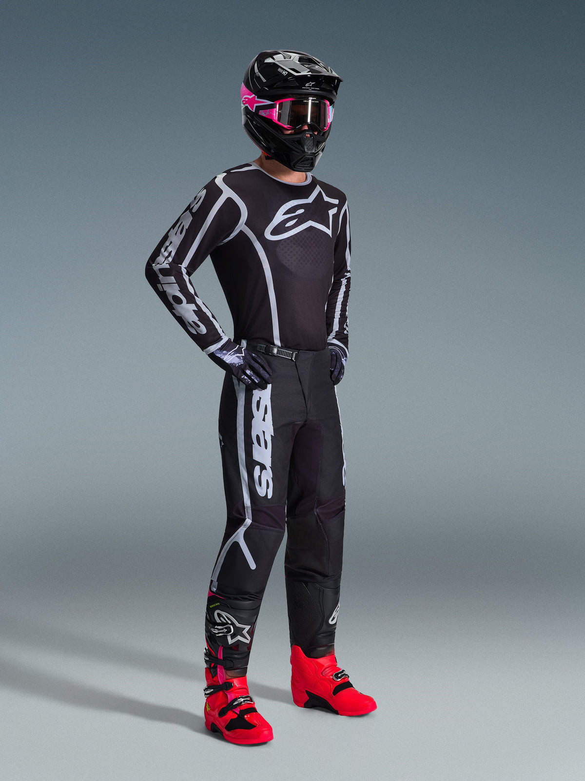 Alpinestars 2026 Fluid Apex Gear Set, pilota di motocross che indossa maglia e pantaloni neri con accenti grafici bianchi e grigi, casco nero con occhiali rosa e stivali da corsa rosa acceso, completo da motocross professionale coordinato