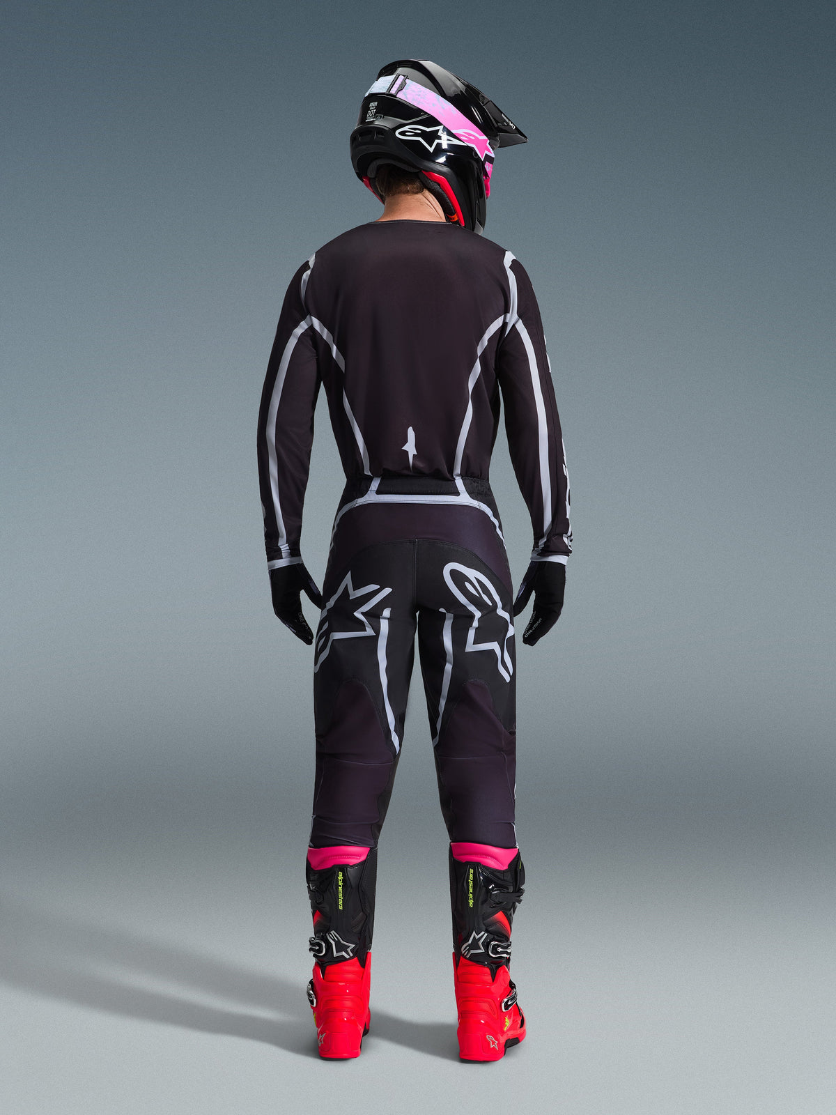 Pilota di motocross visto da dietro che indossa il completo Alpinestars 2026 Fluid Apex, maglia e pantaloni neri con linee geometriche grigie e loghi a stella, casco nero con dettagli rosa, stivali rosso acceso e neri, abbigliamento da gara motocross coordinato
