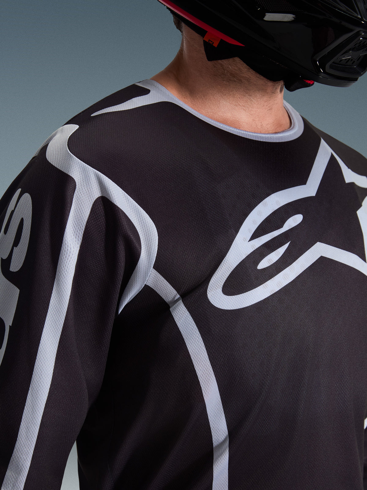 Alpinestars 2026 Fluid Apex Jersey, maglia da motocross, nero e grigio, con grande logo Alpinestars sul petto, colletto sportivo, indossata con un casco nero, abbigliamento tecnico per la guida off-road