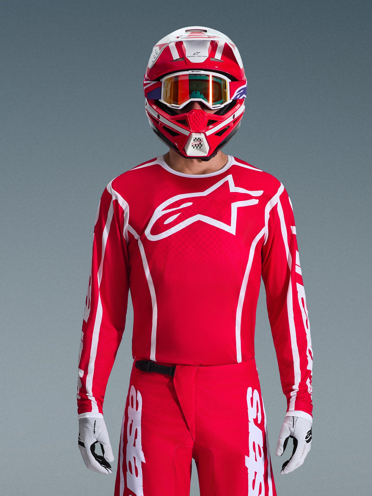 Maglia Alpinestars 2026 Fluid Apex, maglia da motocross, rossa e bianca, indossata da un pilota con casco coordinato rosso e bianco e occhiali arancioni, caratterizzata da un prominente logo bianco sul petto, vestibilità atletica, parte di un set coordinato per corse off-road