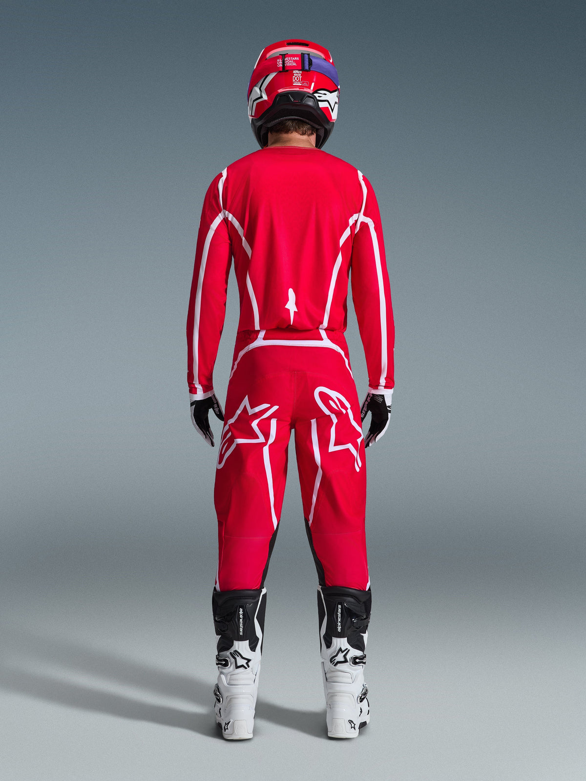 Pilota di motocross visto da dietro che indossa la maglia Alpinestars 2026 Fluid Apex in rosso e bianco, pantaloni rossi coordinati con evidenti loghi a stella bianchi, casco rosso, stivali da motocross bianchi e neri e guanti bianchi e neri, kit completo per gare off-road