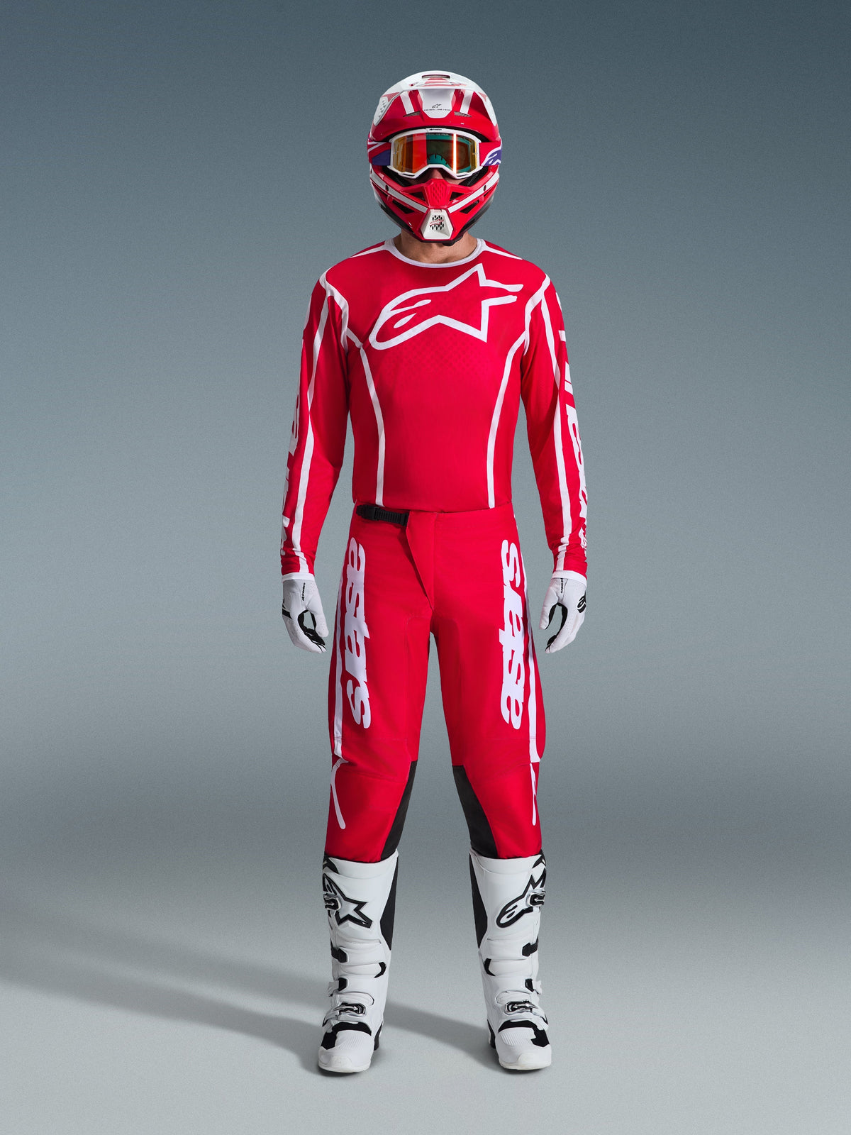 Pilota di motocross con abbigliamento da gara completo Fluid Apex 2026, maglia rossa con logo Alpinestars bianco e branding sulle maniche, pantaloni rossi coordinati con accenti bianchi e pannelli neri, casco rosso e bianco con occhiali a specchio, stivali bianchi e neri e guanti bianchi e neri, set professionale per prestazioni fuoristrada.