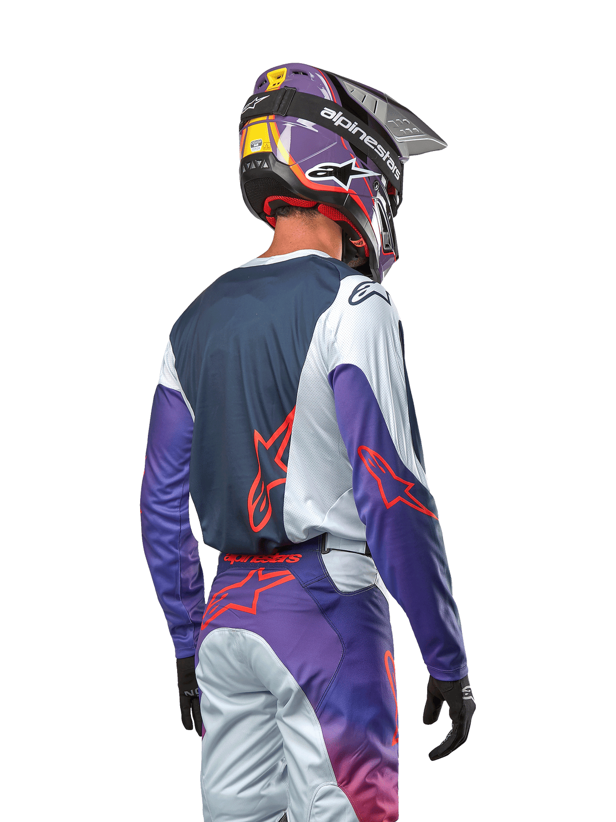 Pilota di motocross visto di spalle con indosso la maglia Alpinestars Racer Hoen 2024, pannello posteriore blu navy con spalle grigio chiaro e maniche viola, loghi Alpinestars arancione acceso, pantaloni coordinati viola e grigi e casco viola, kit da gara off-road completo
