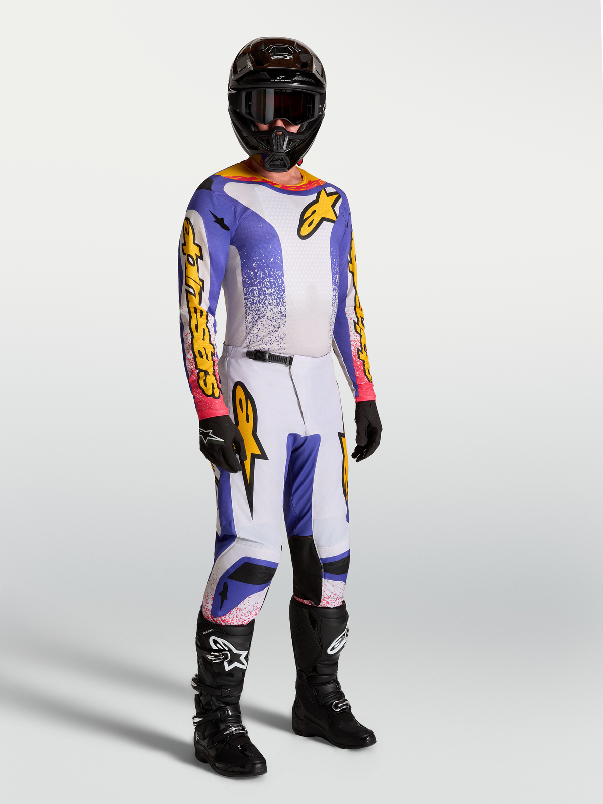 Alpinestars Fluid Drag Gear Set e pantaloni Fluid Drag, completo e pantaloni motocross da uomo, lilla, blu iris e oro, indossati da un modello con casco e stivali neri, abbigliamento performance per guida off-road.