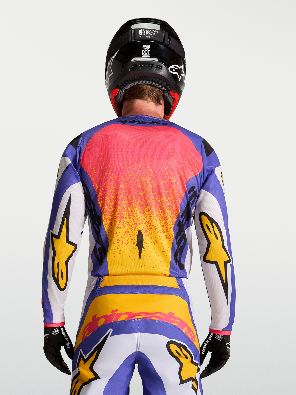 Alpinestars Maglia Fluid Drag, maglia motocross da uomo, colore lilla chiaro e blu con dettagli dorati, vista posteriore su modello con pantaloni e casco coordinati.